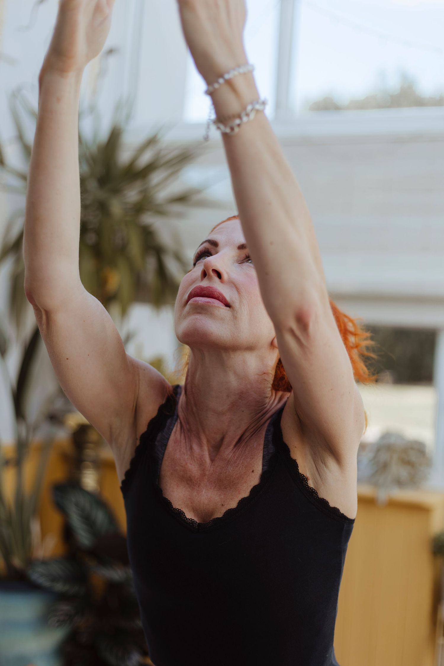 A woman with red hair lifts her arms overhead, appearing focused and engaged in a stretching or yoga movement.