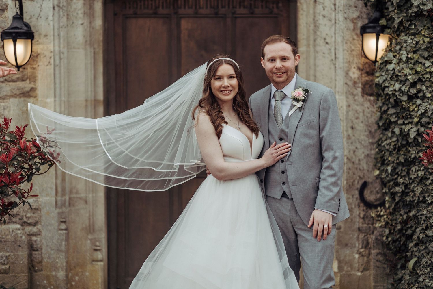 A smiling bride and groom pose together, her veil flowing as they stand in front of a rustic doorway.