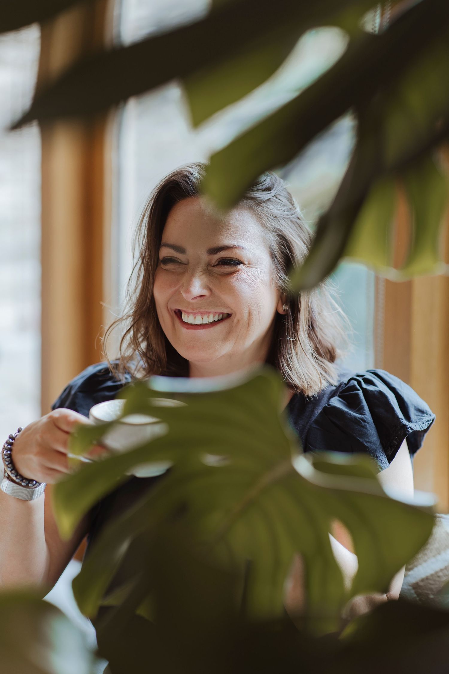 A woman smiles warmly while enjoying a drink, framed by soft, leafy greenery in a bright, peaceful space.