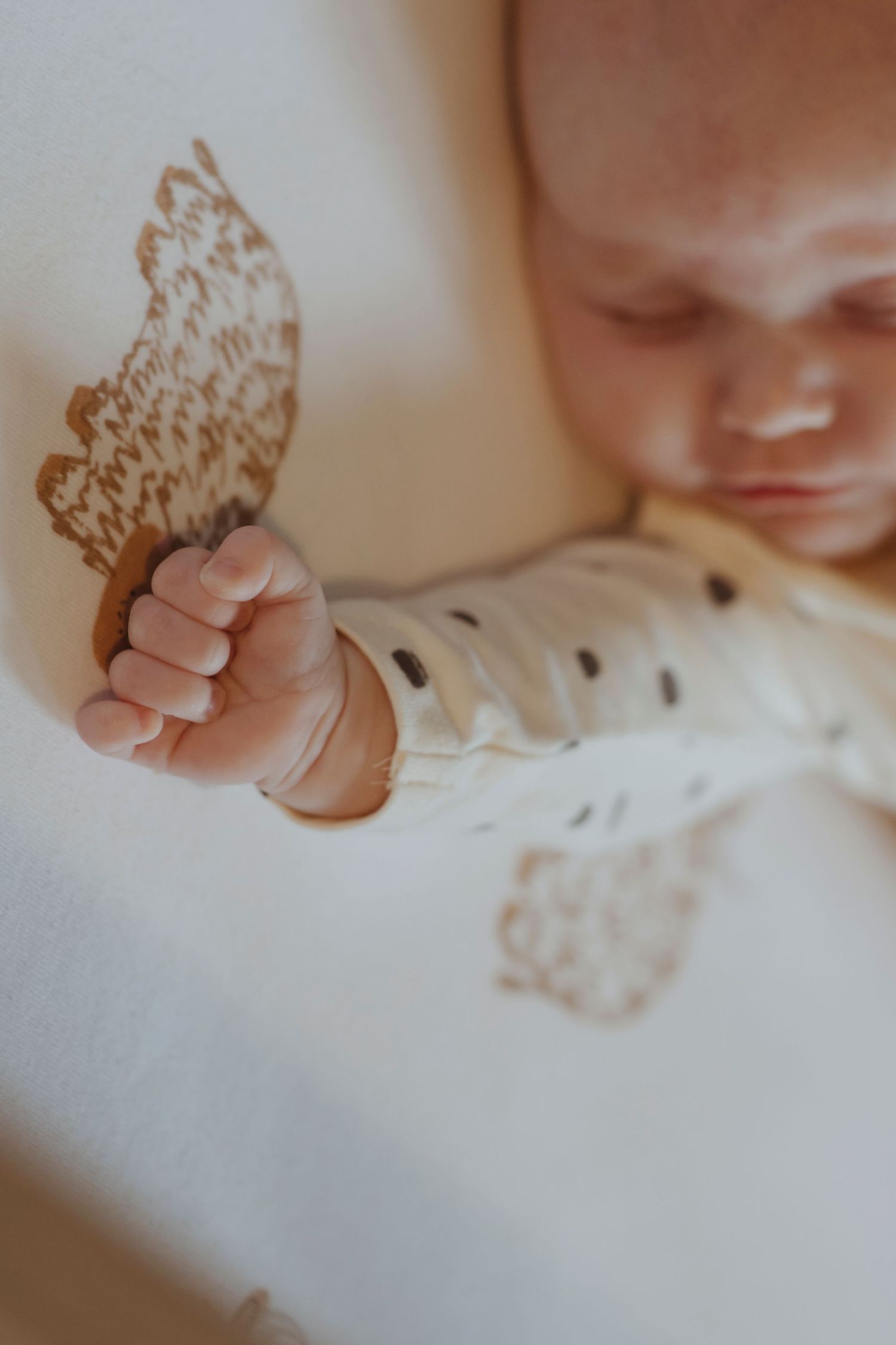 A baby sleeps with a tiny fist resting near a soft, wing-shaped design on the blanket.