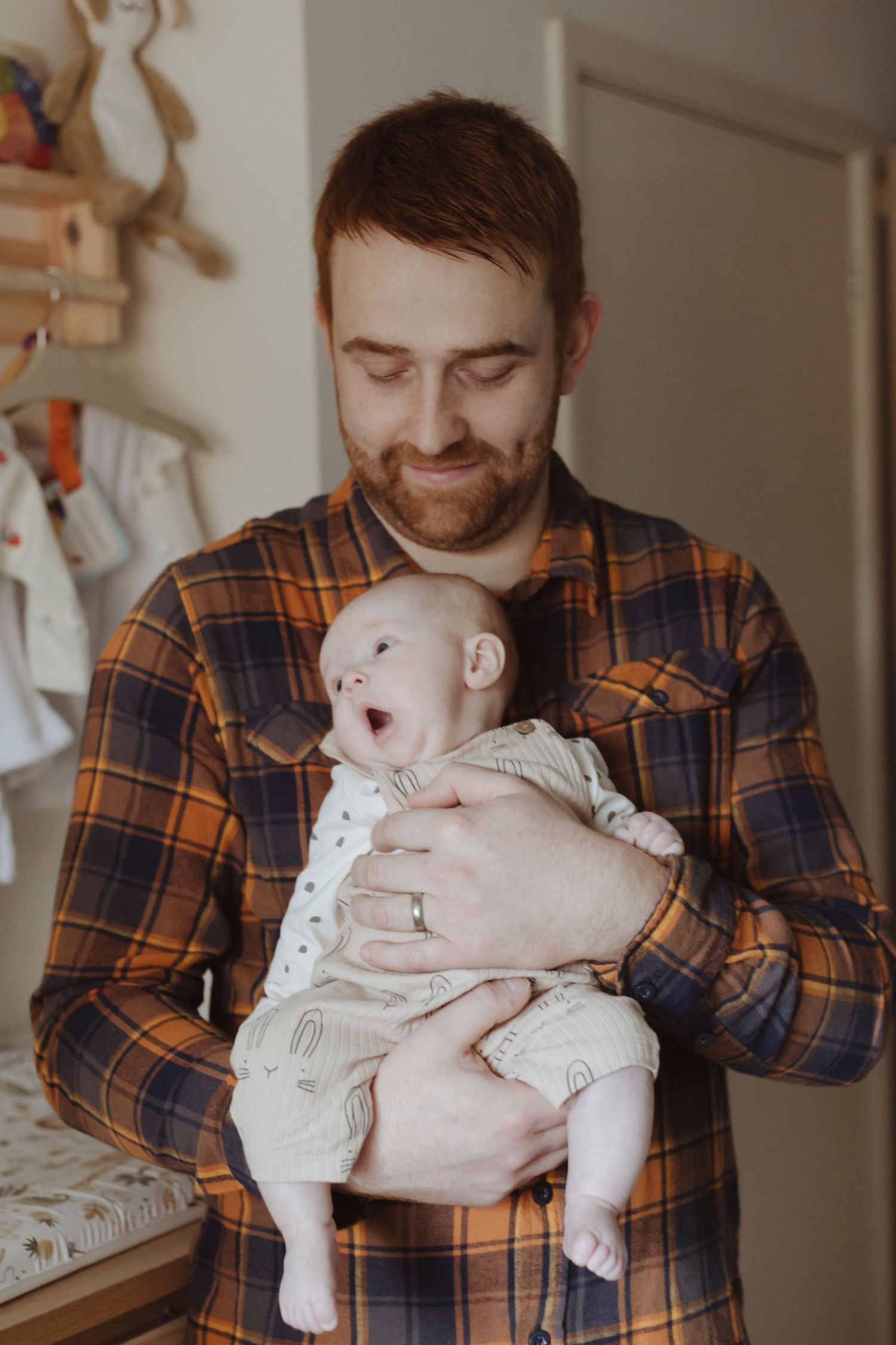 A man holds a baby close, smiling softly as the baby looks up with an open-mouthed expression.