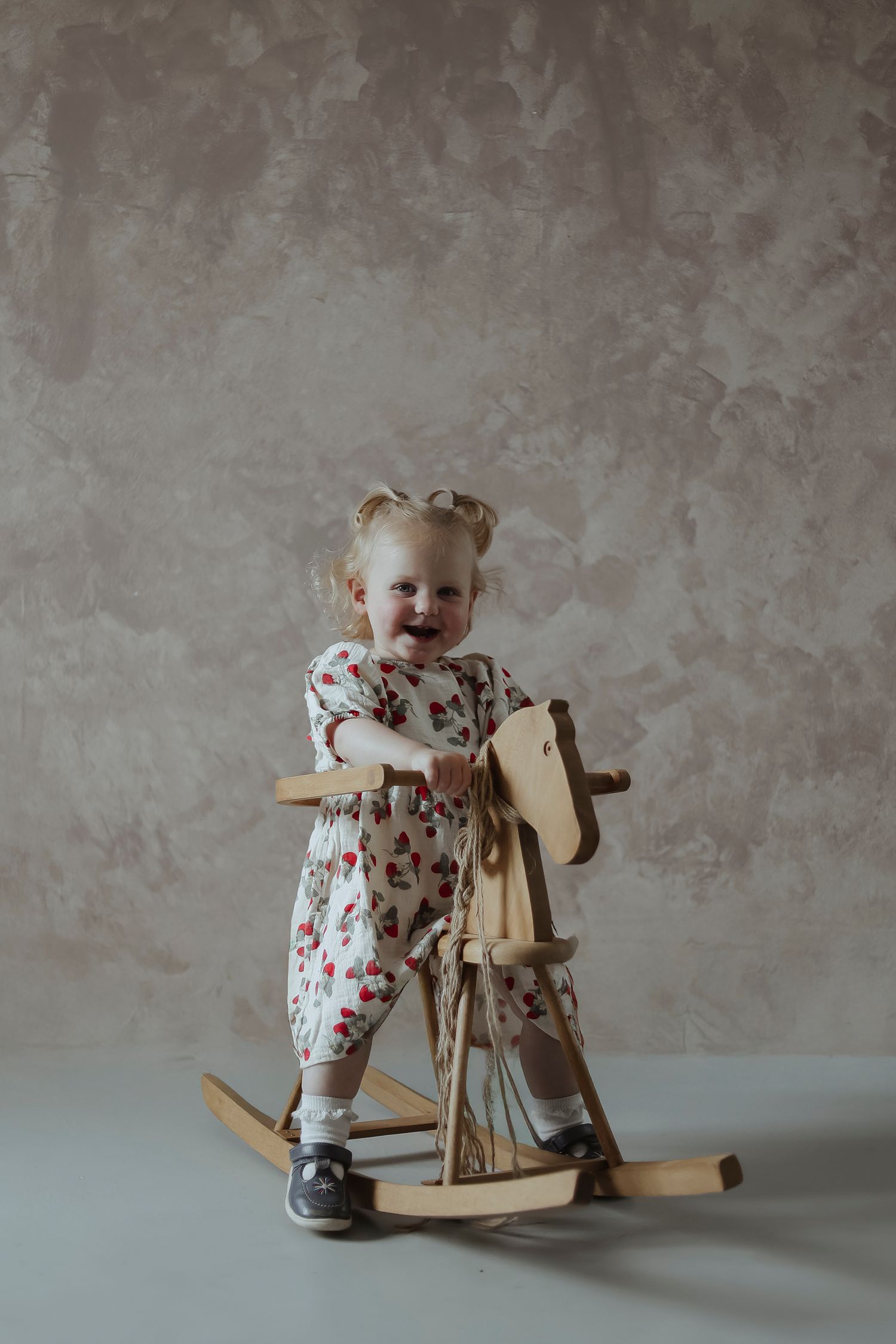 A small child in patterned clothes smiles while holding onto a wooden rocking toy in a simple studio setting.