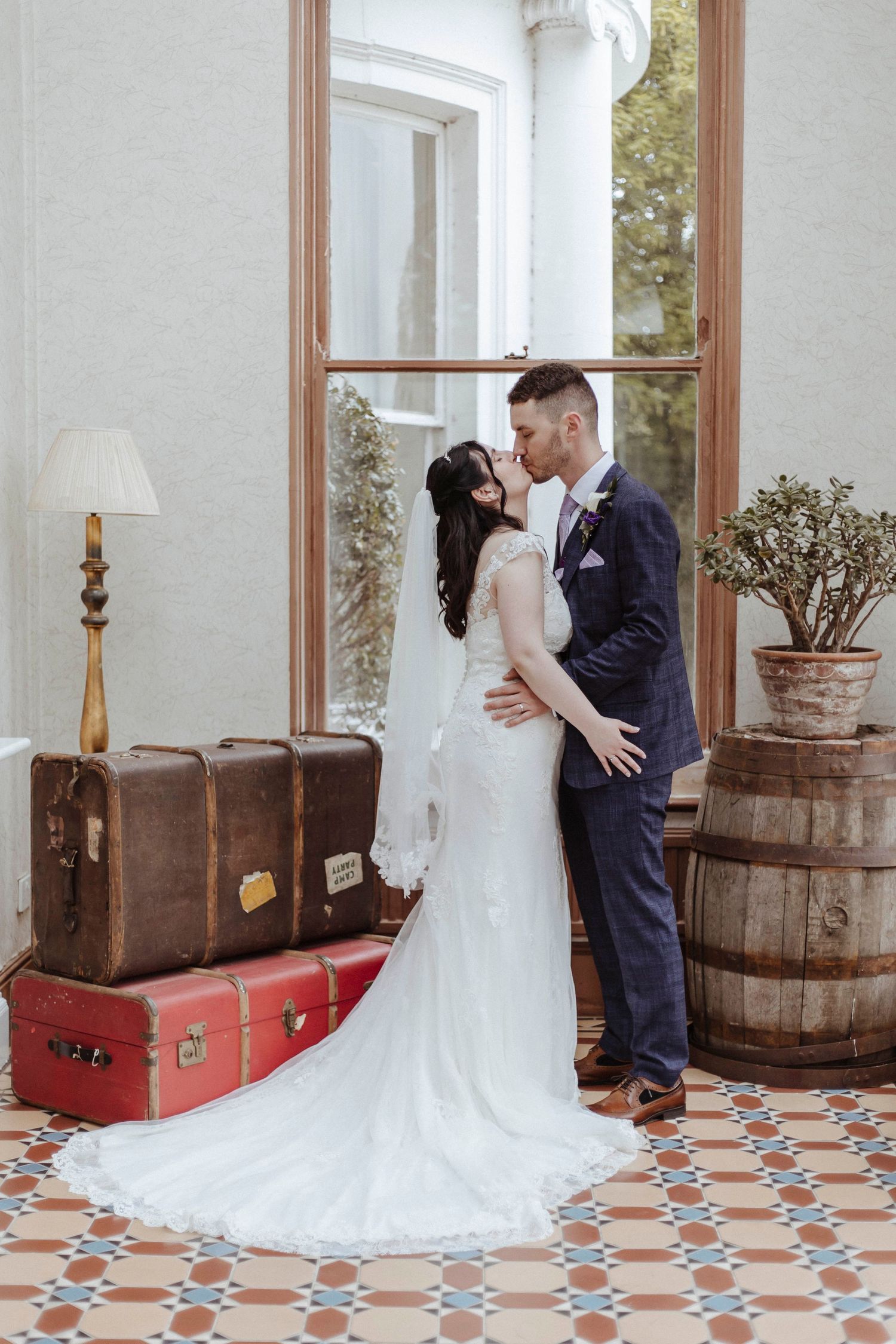 A bride and groom share a kiss indoors beside stacked suitcases and a wooden barrel.
