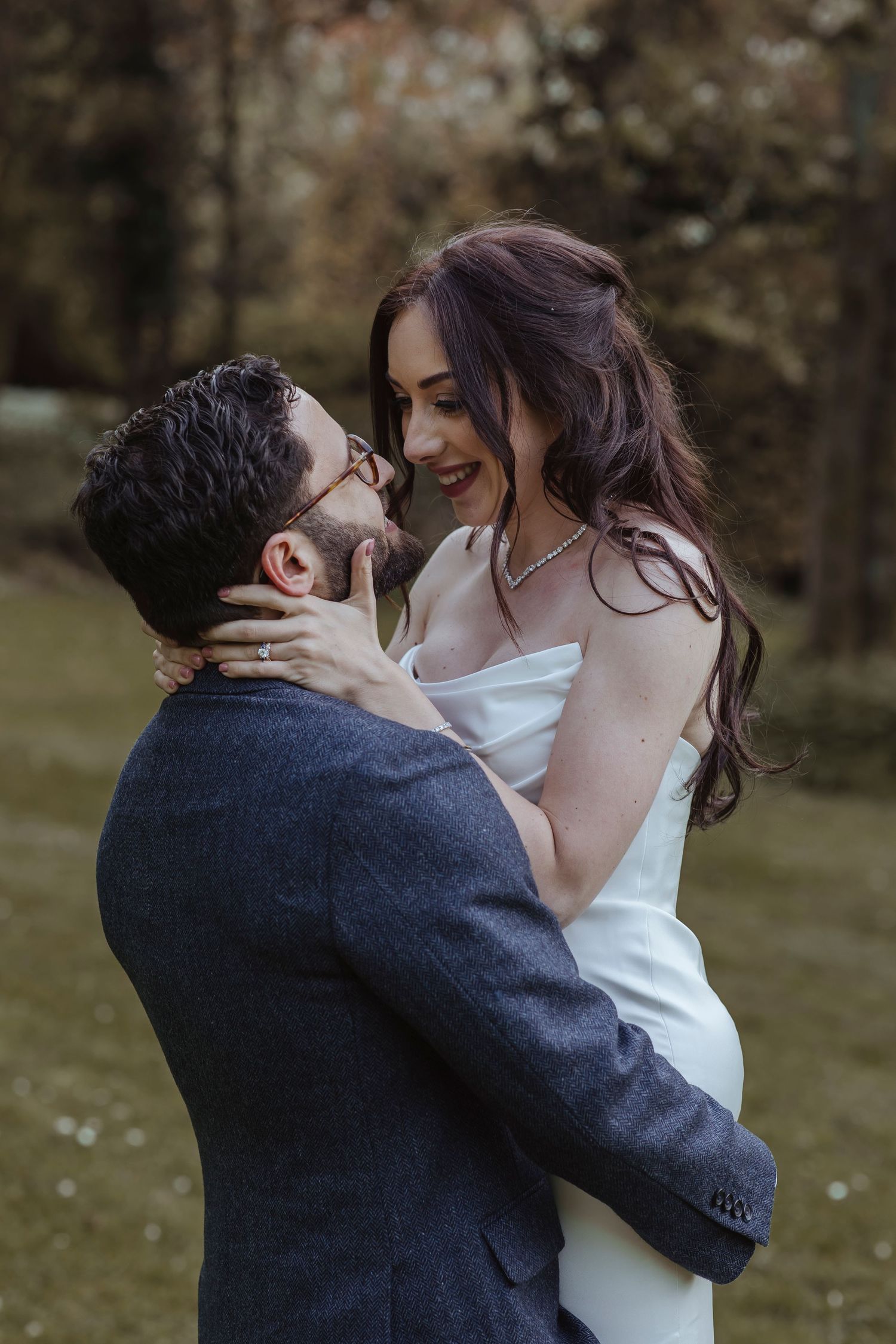 A couple embraces outdoors, smiling closely at each other as he lifts her and she holds his shoulders in a warm, romantic moment.