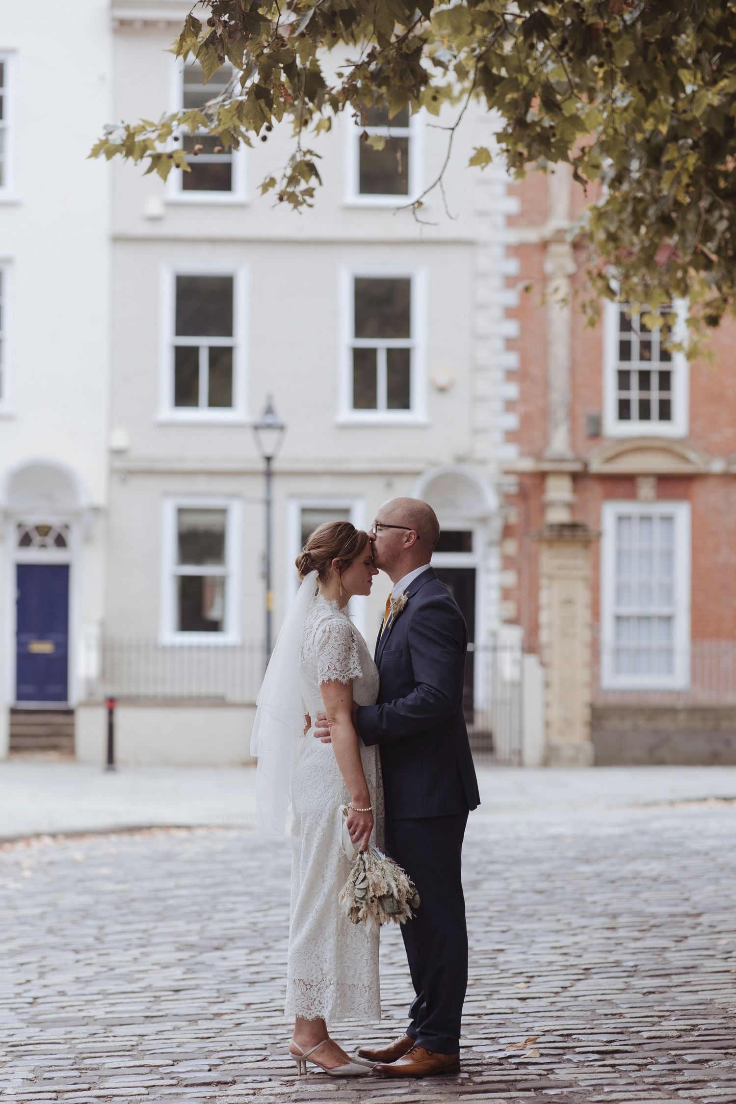 A couple stands close on a cobblestone street, gently touching foreheads as they share a quiet, intimate moment.