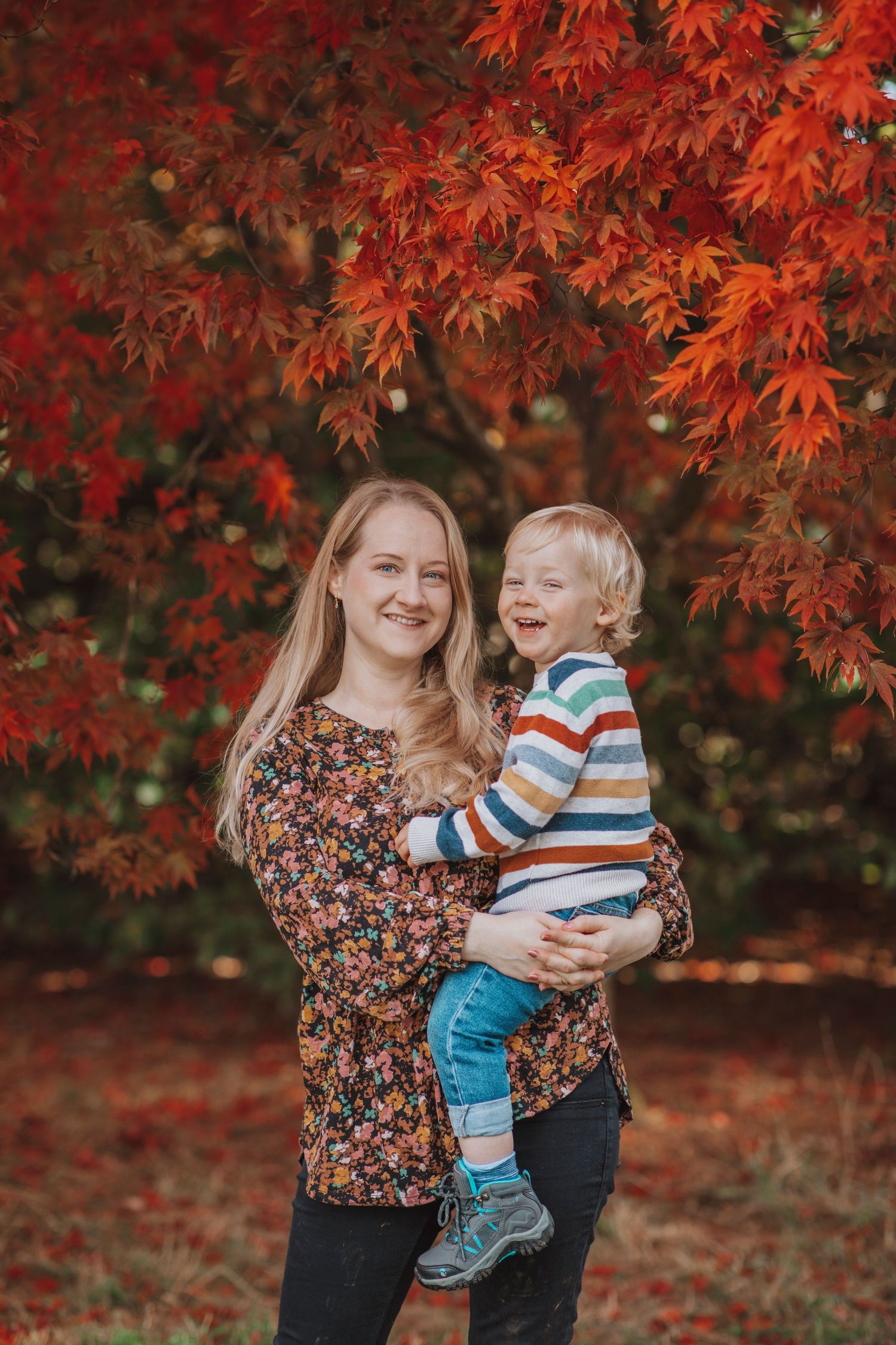 A woman holding a smiling child stands under vibrant red autumn leaves in an outdoor setting.
