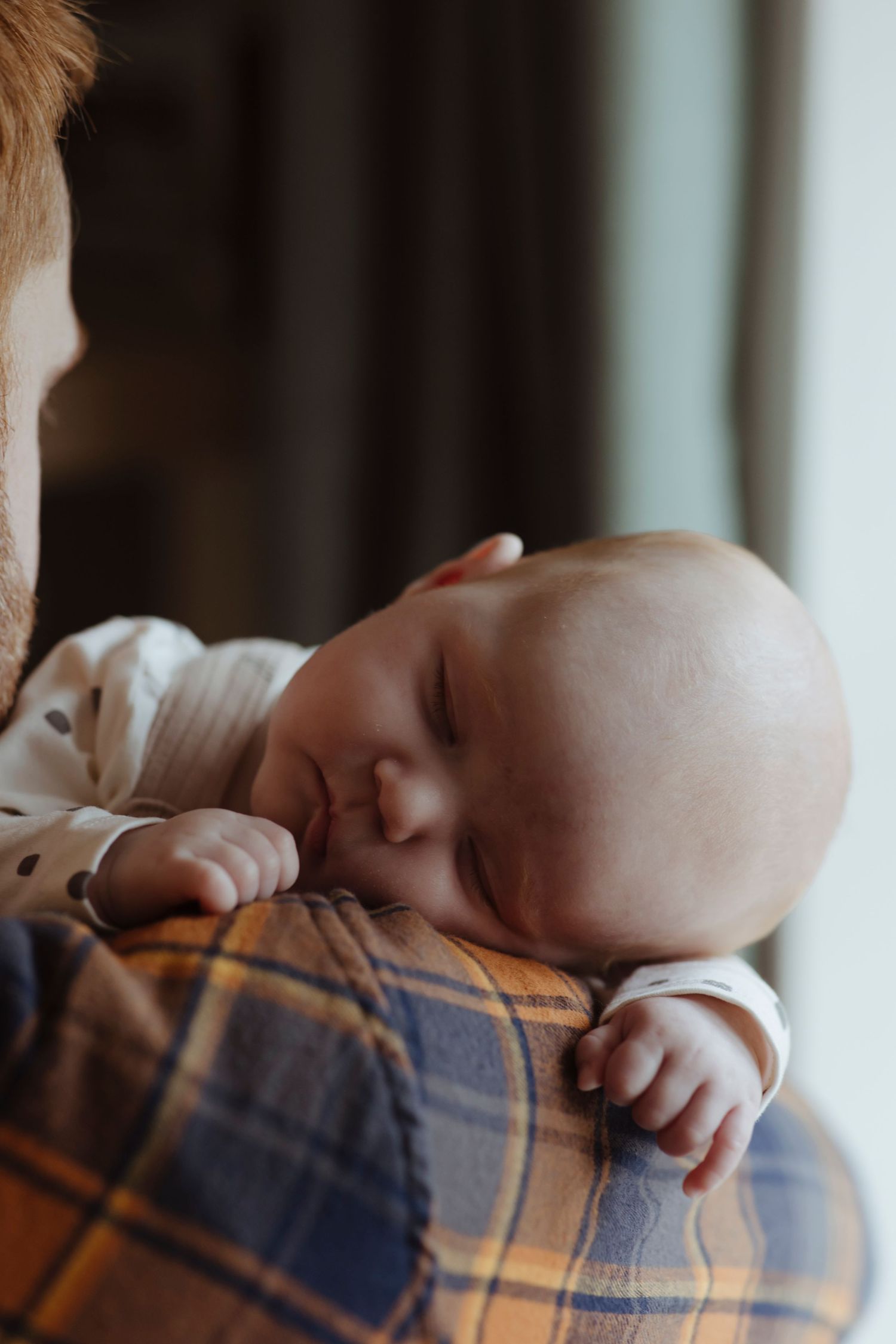 A sleeping baby rests on an adult’s shoulder, nestled peacefully with a tiny hand draped over the person’s plaid shirt.