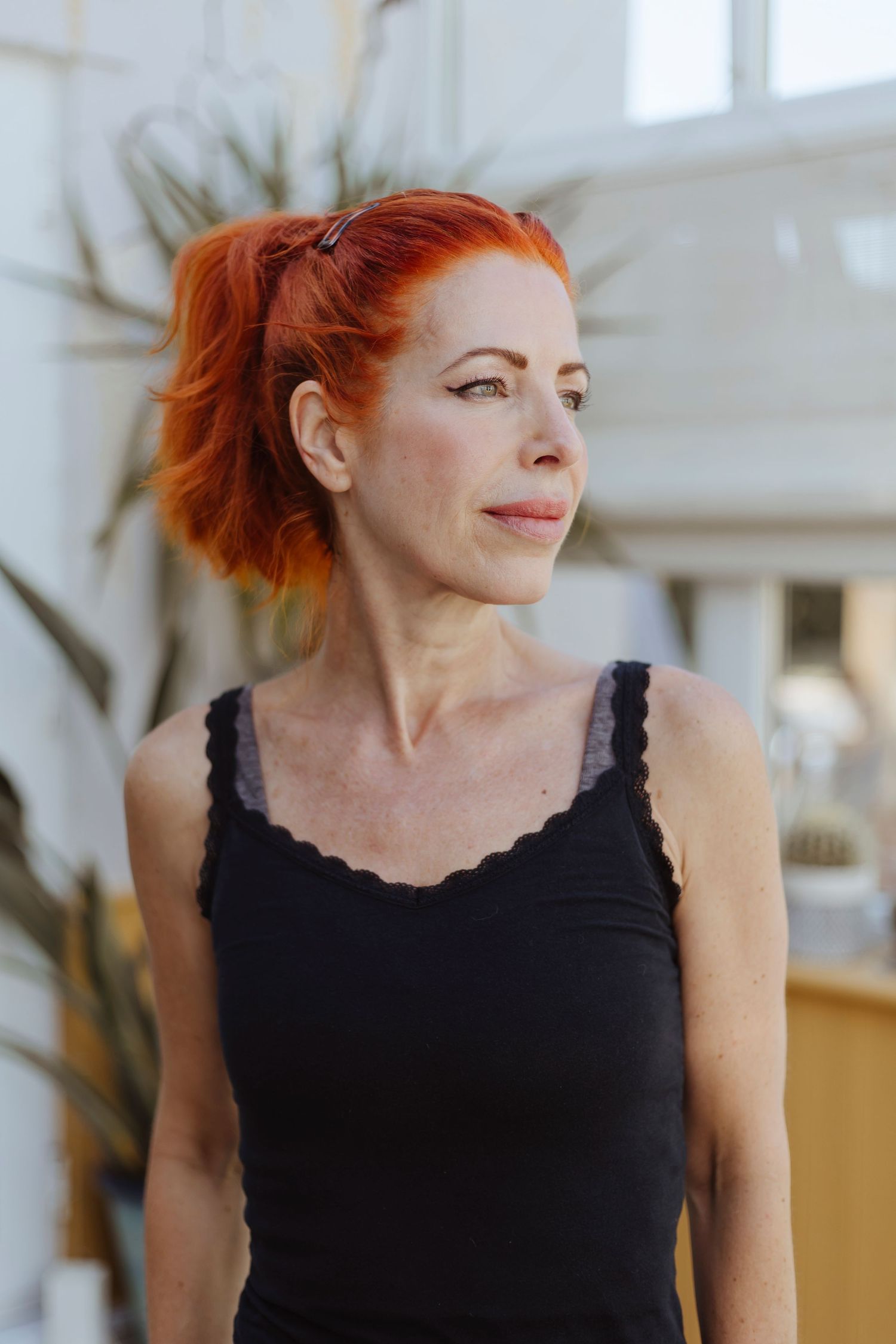 A woman with bright red hair stands indoors, looking to the side with a calm, reflective expression.
