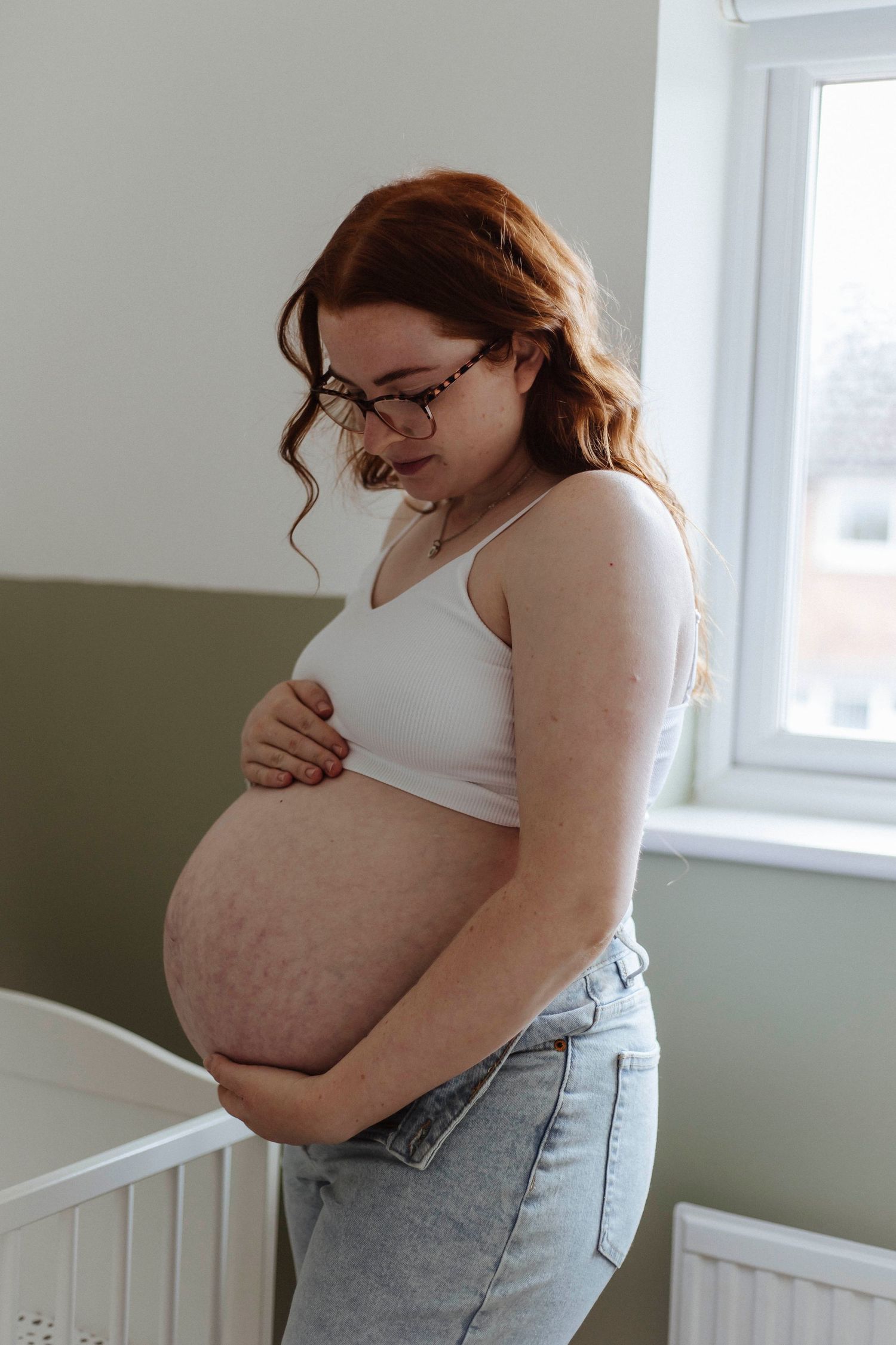 A quiet, tender moment as a pregnant woman cradles her belly, standing softly in the nursery light.