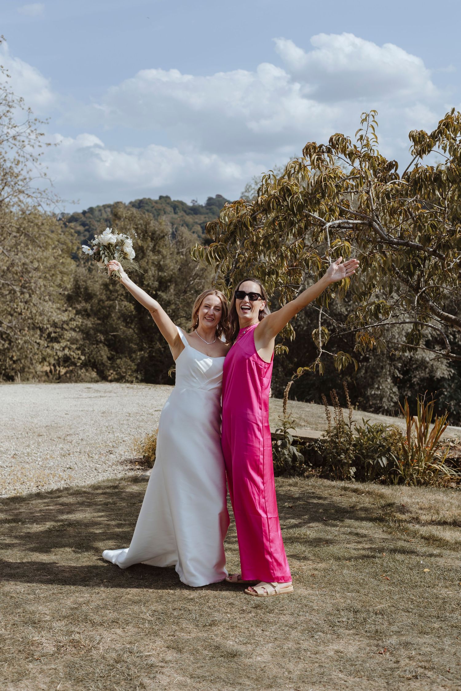 A joyful bride and her friend pose with arms raised, celebrating outdoors under bright skies and surrounded by nature.