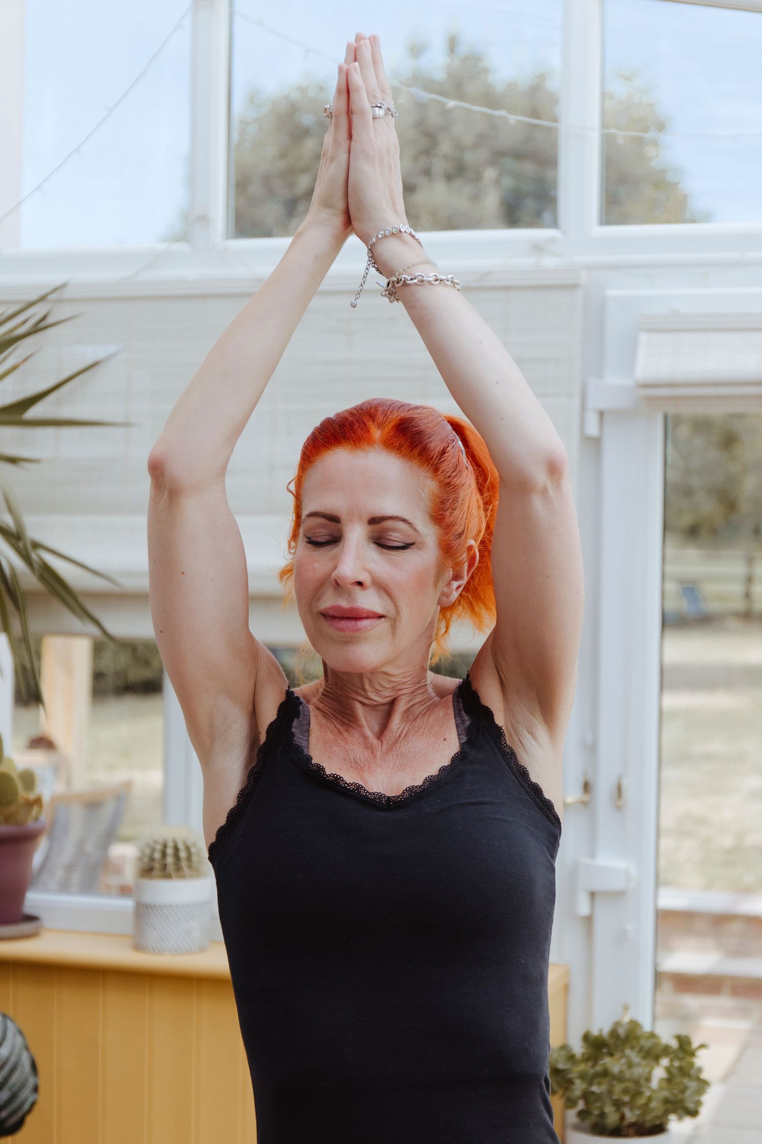A woman with red hair stands indoors in a yoga pose, eyes closed and hands pressed together overhead, looking calm and centered.