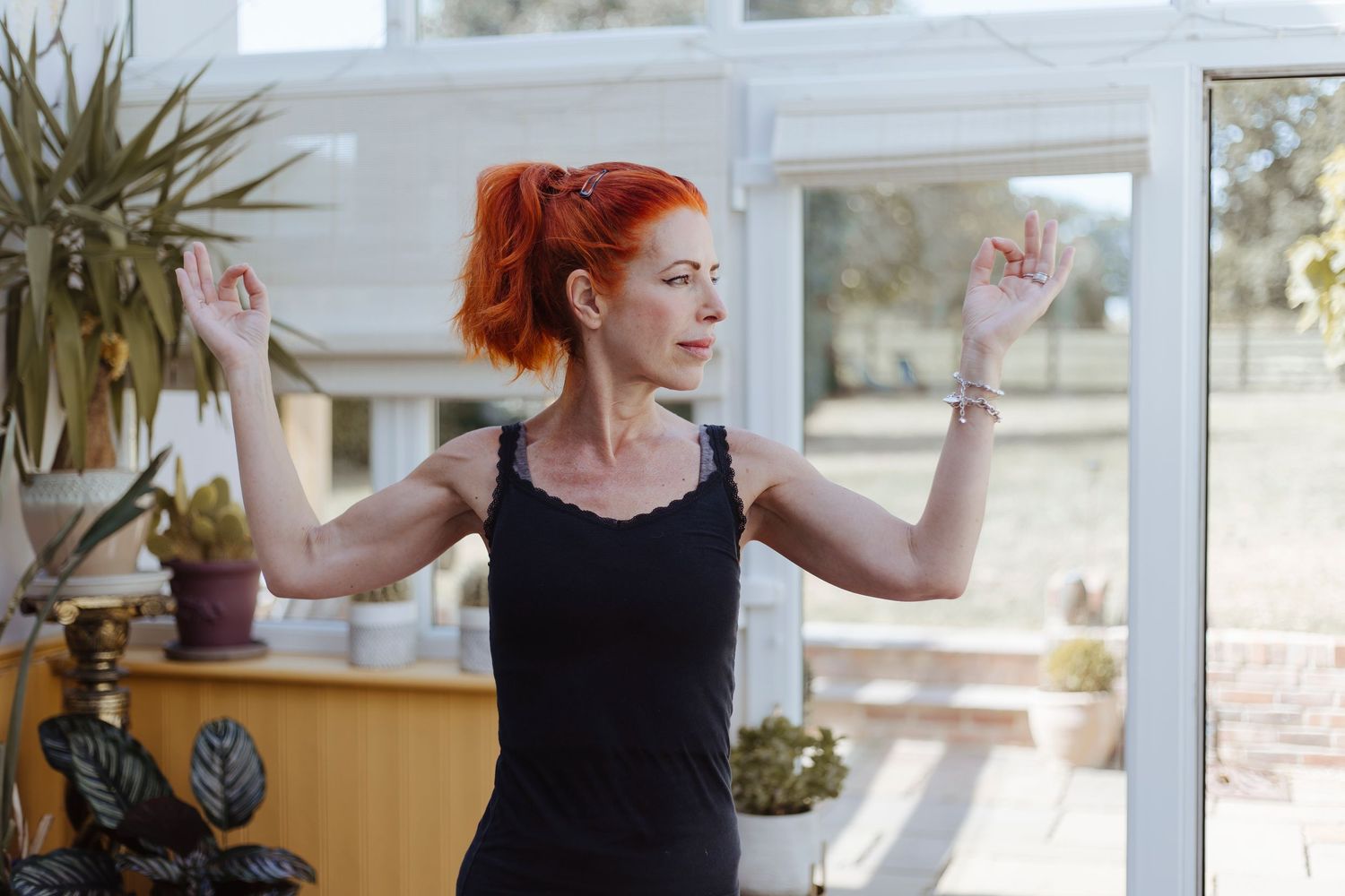 A person stands in a bright room with plants, arms raised as if stretching or moving gracefully, looking calmly to the side.
