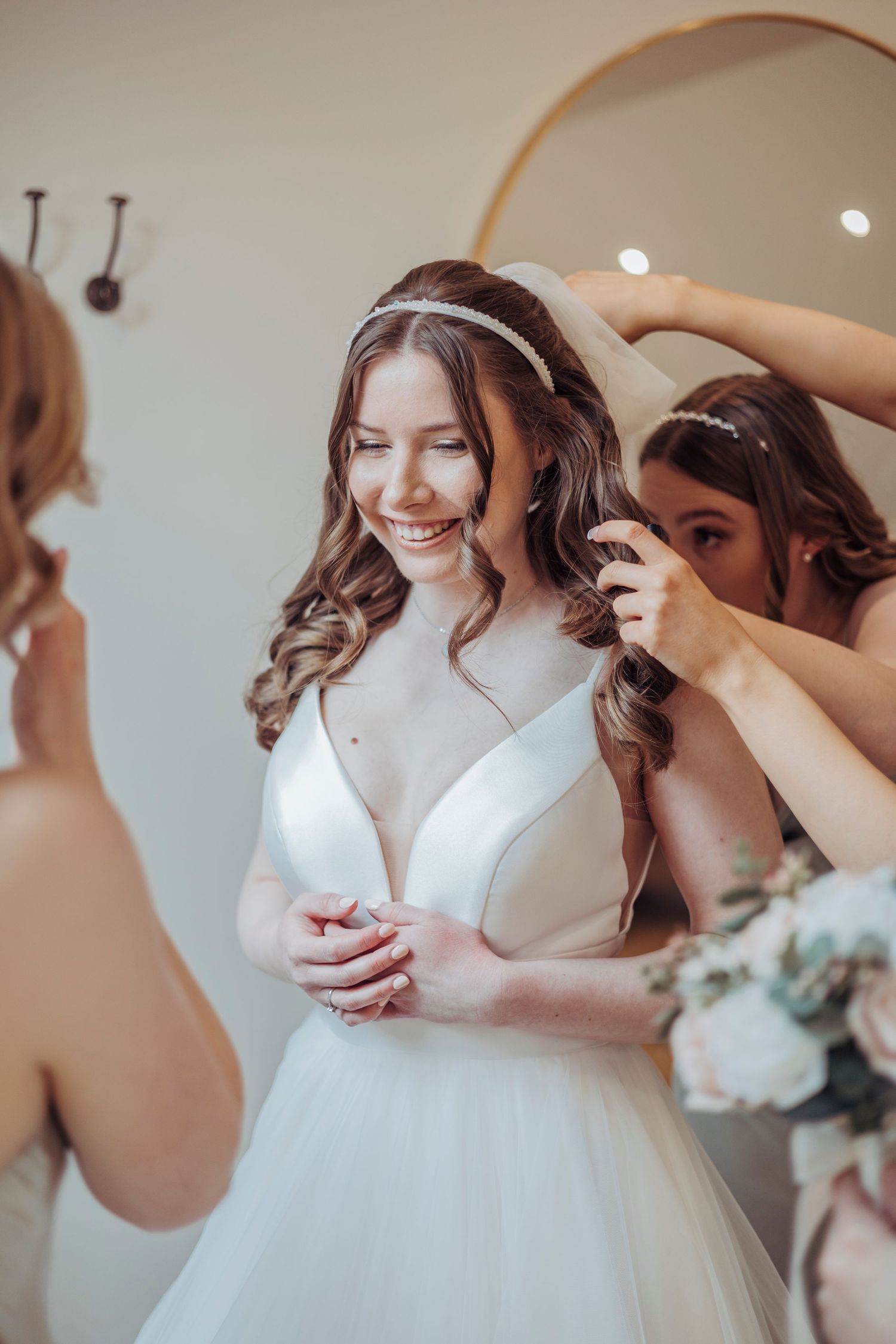 A radiant bride gets her veil adjusted by a bridesmaid, smiling softly as she prepares for the ceremony.