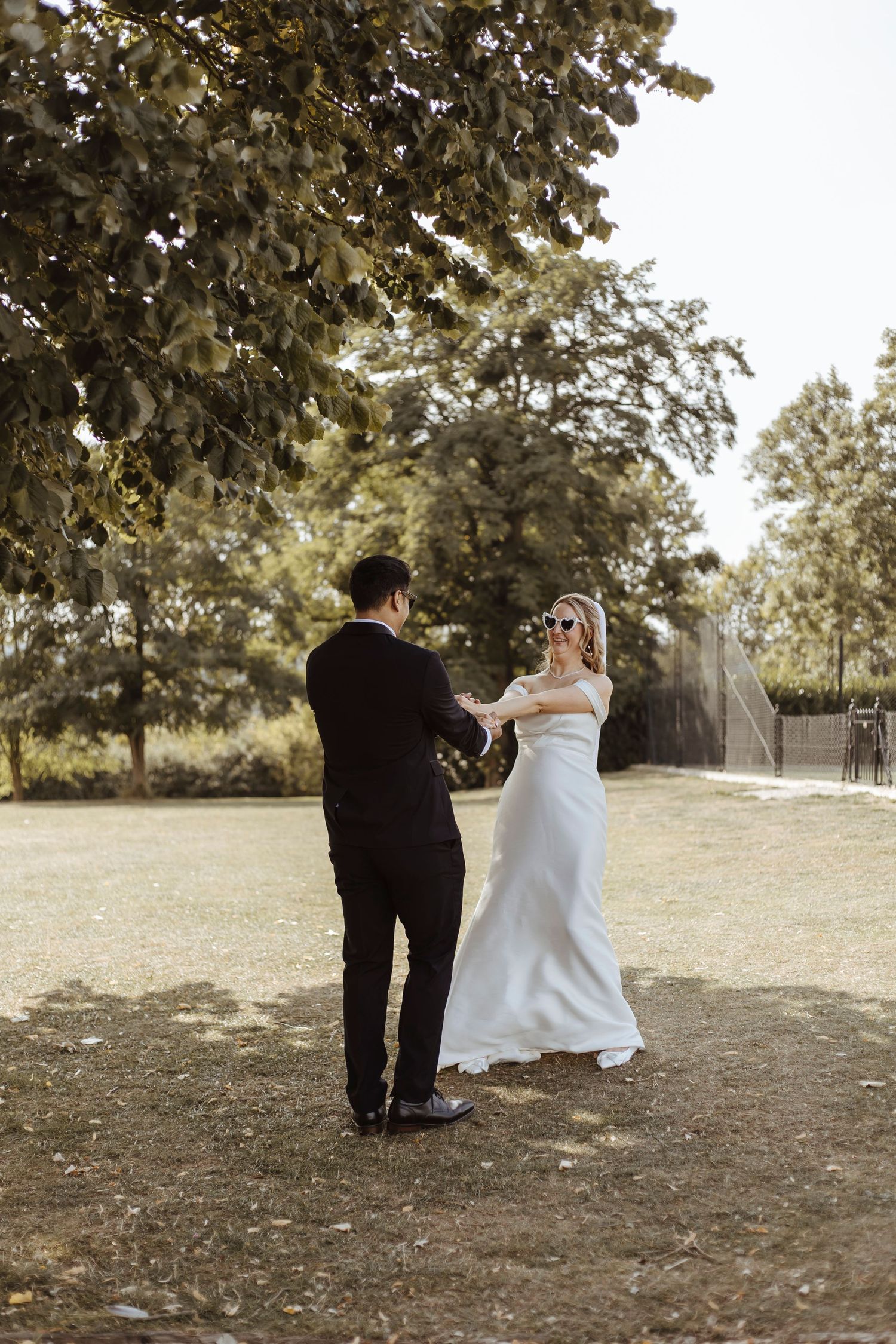 A bride and groom share a joyful dance beneath sunlit trees.