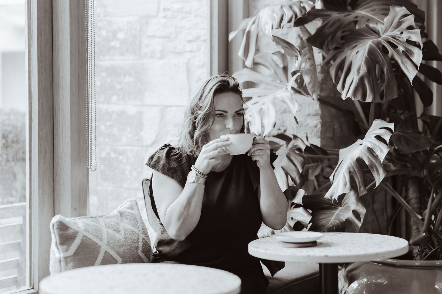A woman enjoys a quiet moment with a warm drink, seated by large windows and lush plants in a calm, cozy café setting.