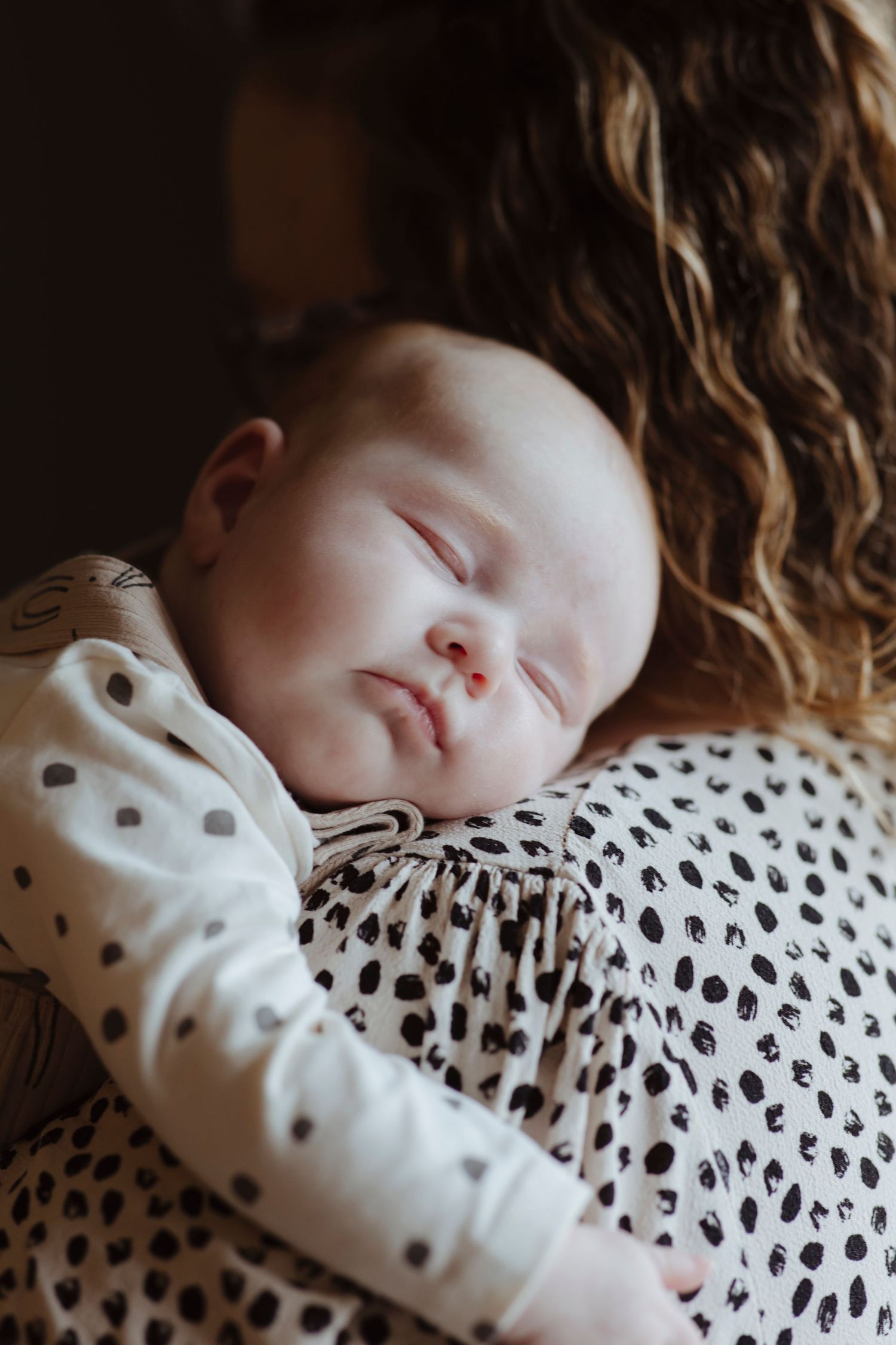 A baby sleeps peacefully on an adult’s shoulder, resting against a patterned shirt.