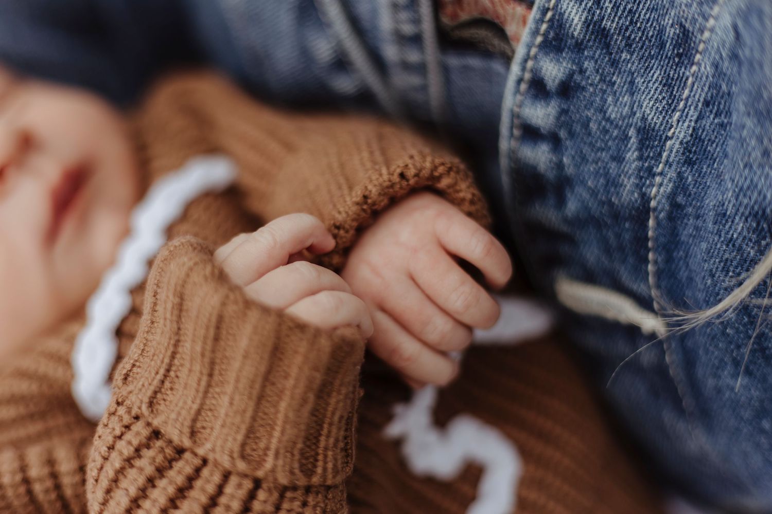 A tiny hand peeks from a cozy brown sweater, resting softly against denim in a peaceful, close-up moment.