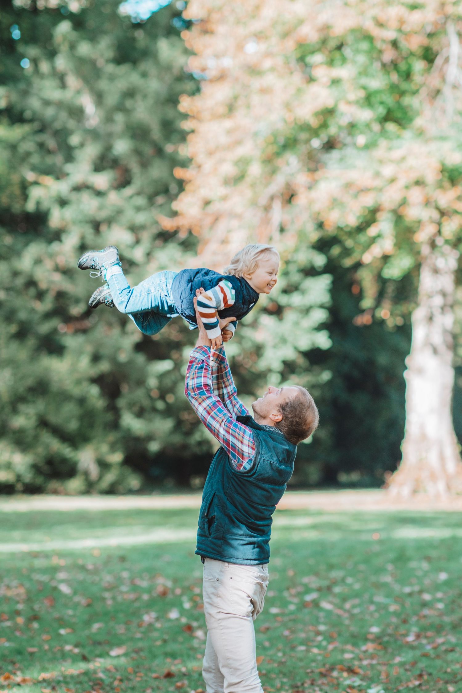 An adult lifts a laughing child high in the air in a sunny park surrounded by tall green trees.