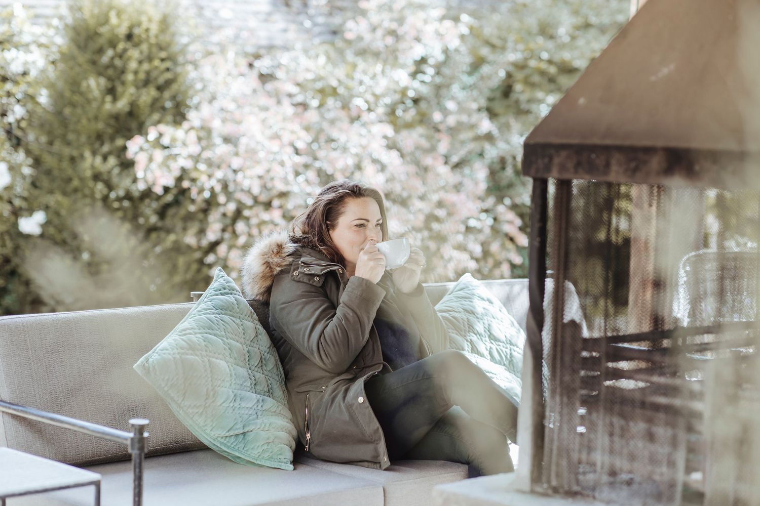 A person relaxes outdoors on a cushioned sofa, sipping a warm drink beside a rustic outdoor fireplace.
