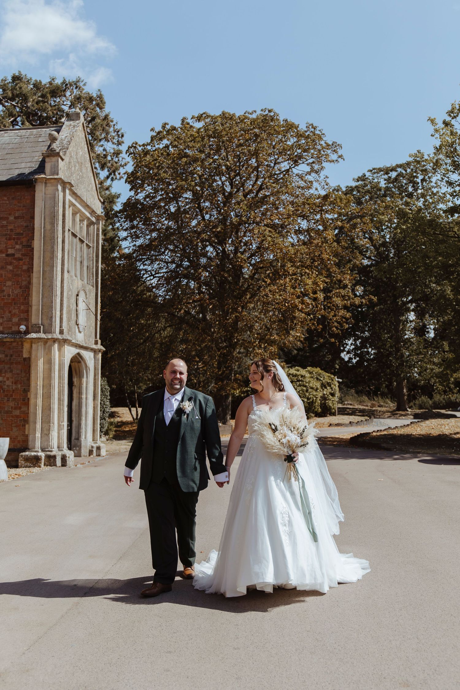 A newly married couple walks hand in hand outdoors, dressed in their wedding attire, enjoying a bright and sunny moment together.