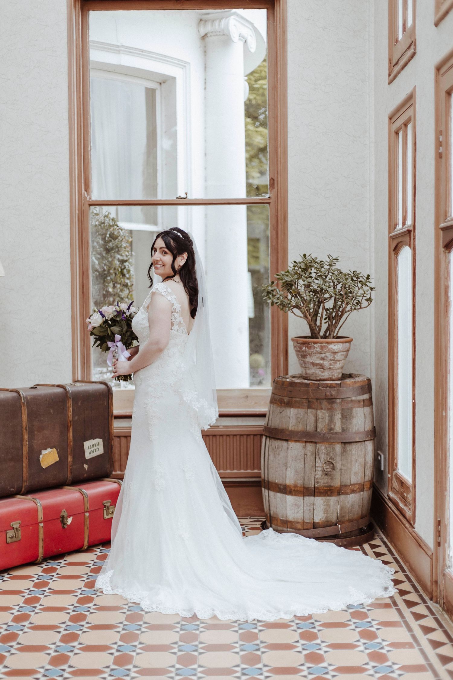 A bride in a long white gown poses indoors beside vintage suitcases and a wooden barrel.