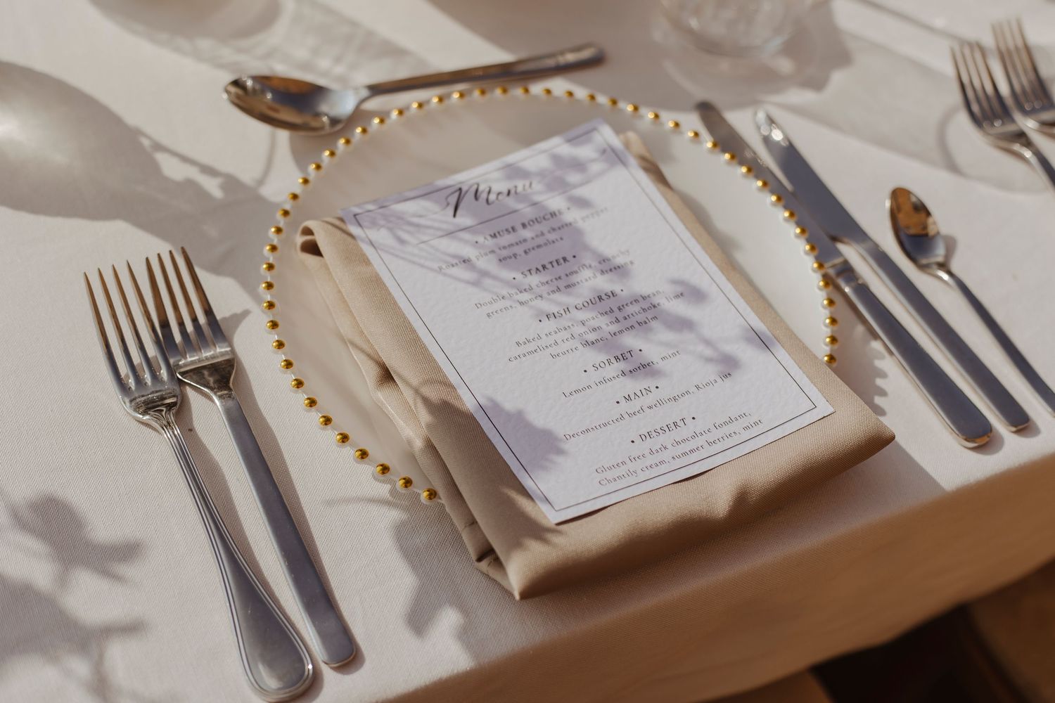 A neatly set table with silverware, a beaded charger, and a folded napkin topped with a printed menu in soft sunlight.