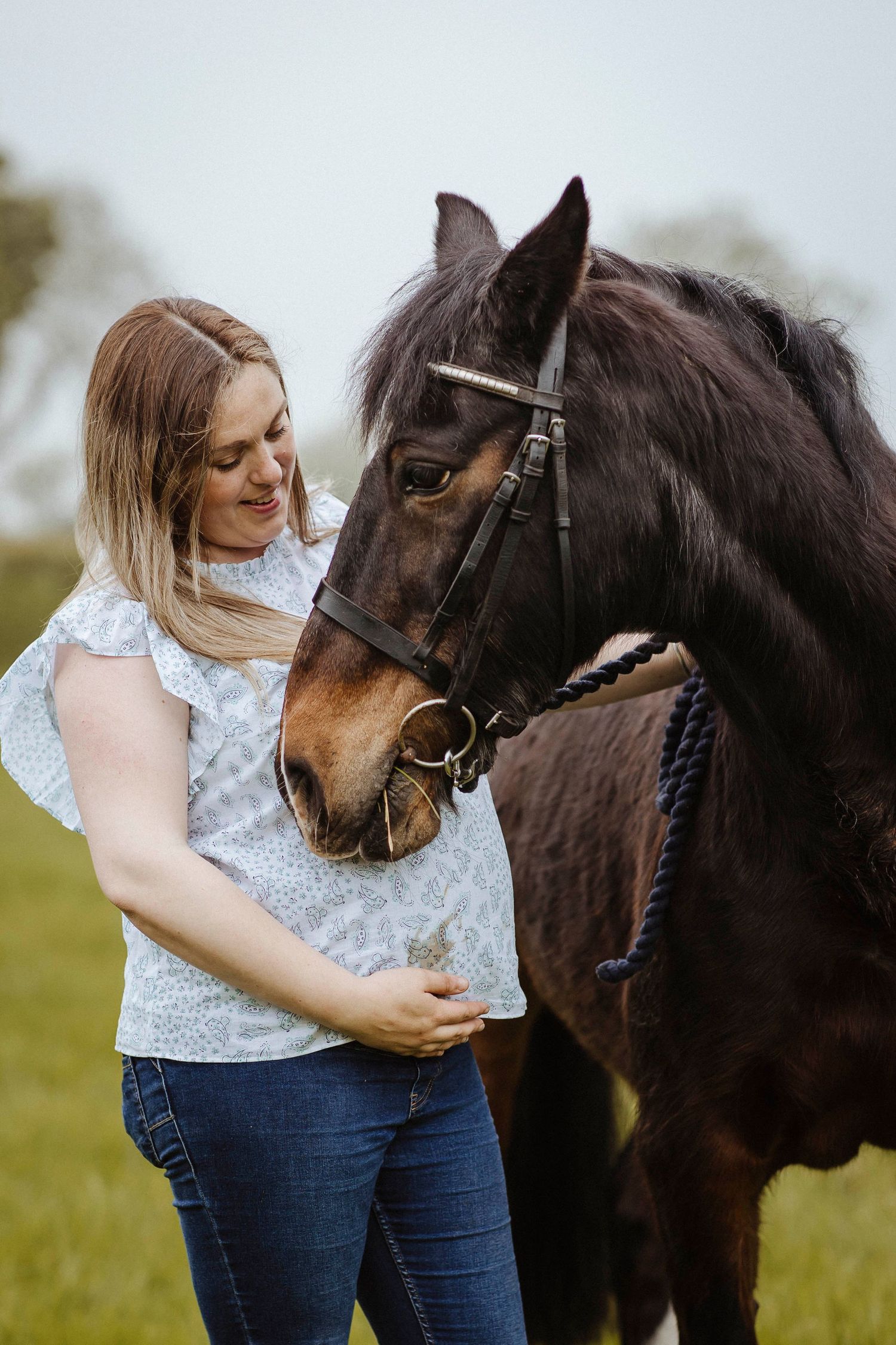 A woman gently rests a hand on her belly while standing close to a calm horse, sharing a warm, tender moment outdoors.