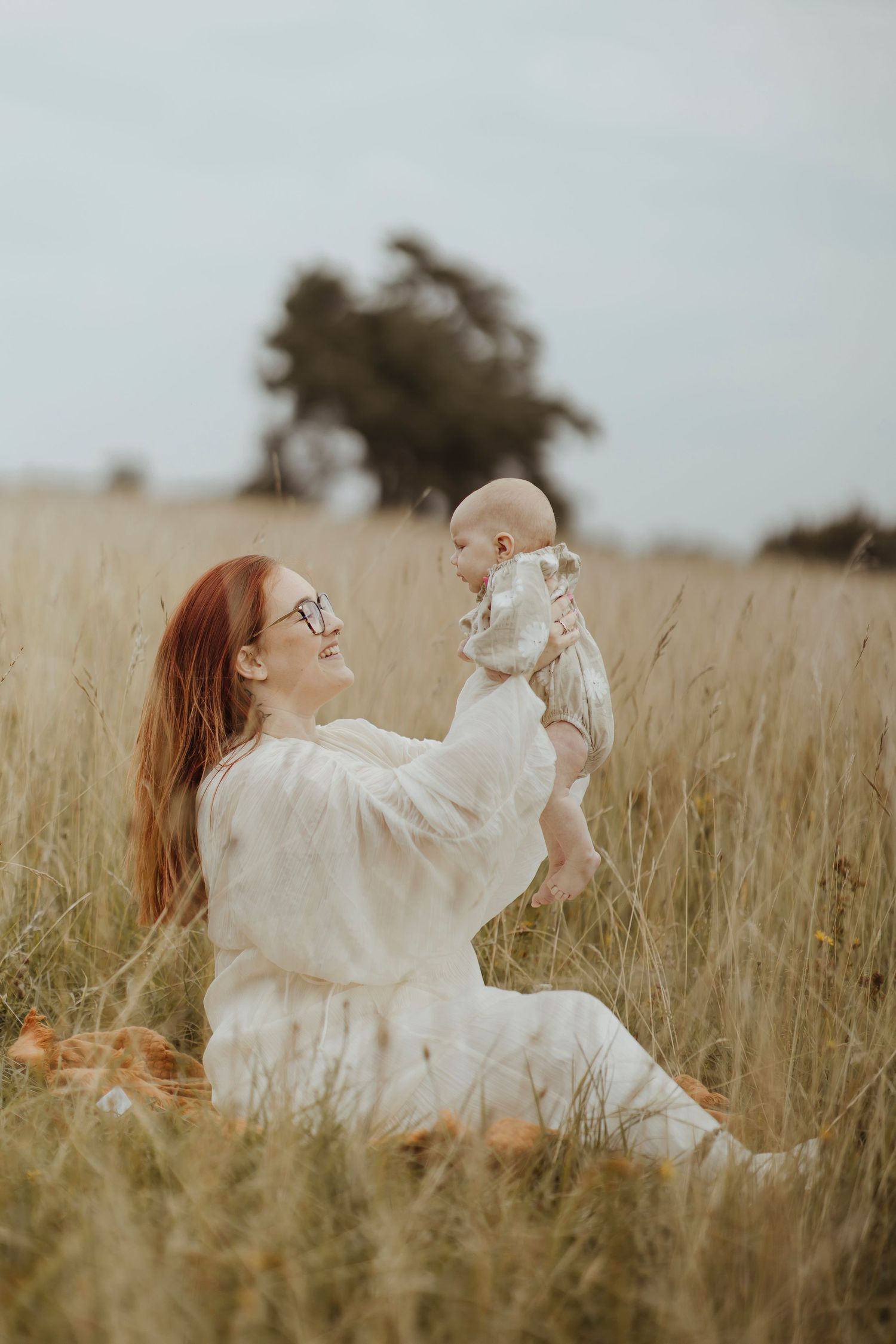 A woman with red hair sits in a tall grassy field, holding a baby up playfully as they look at each other.