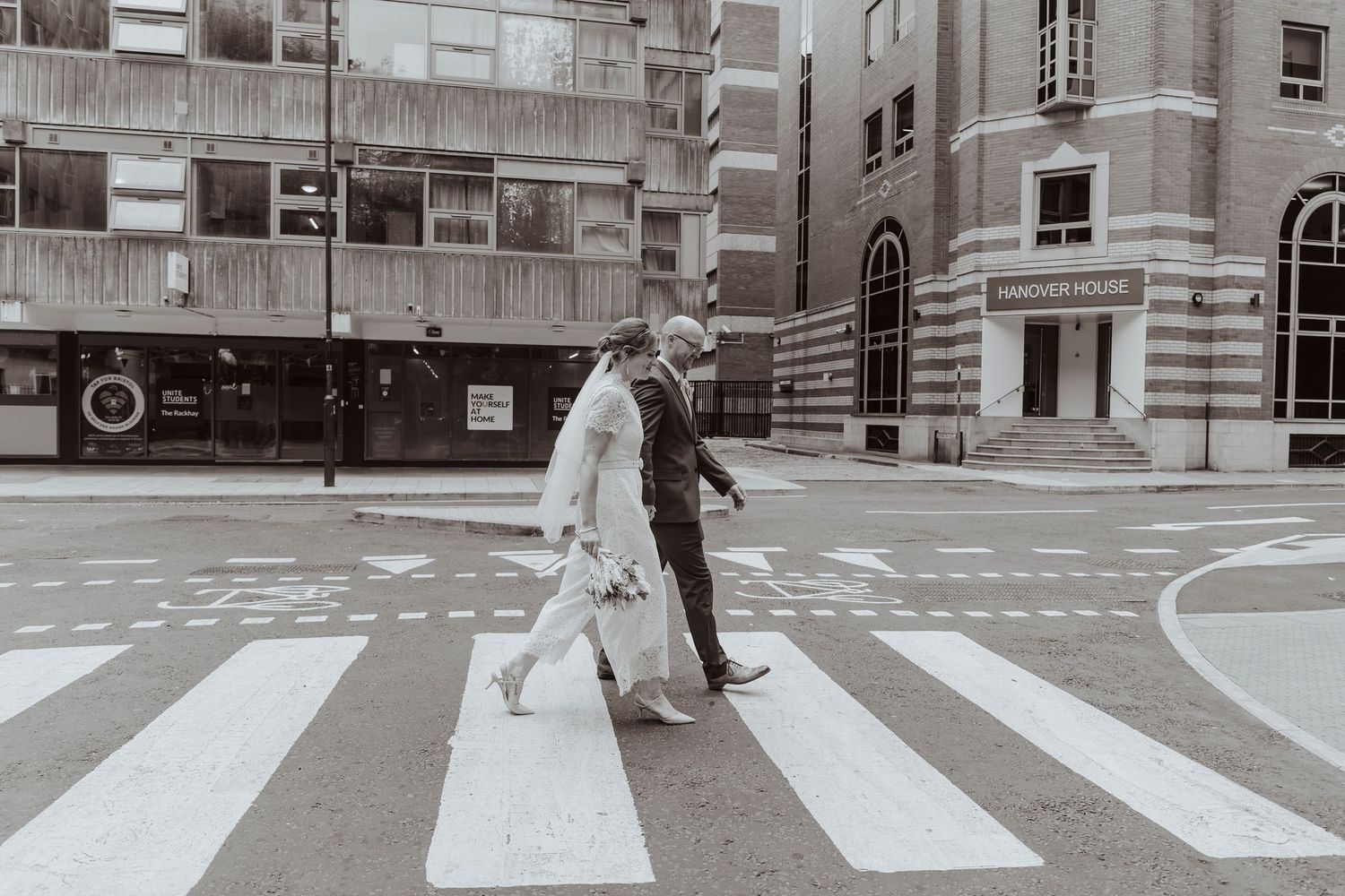 A newlywed couple walks hand in hand across a city crosswalk, captured in a candid, documentary-style moment.