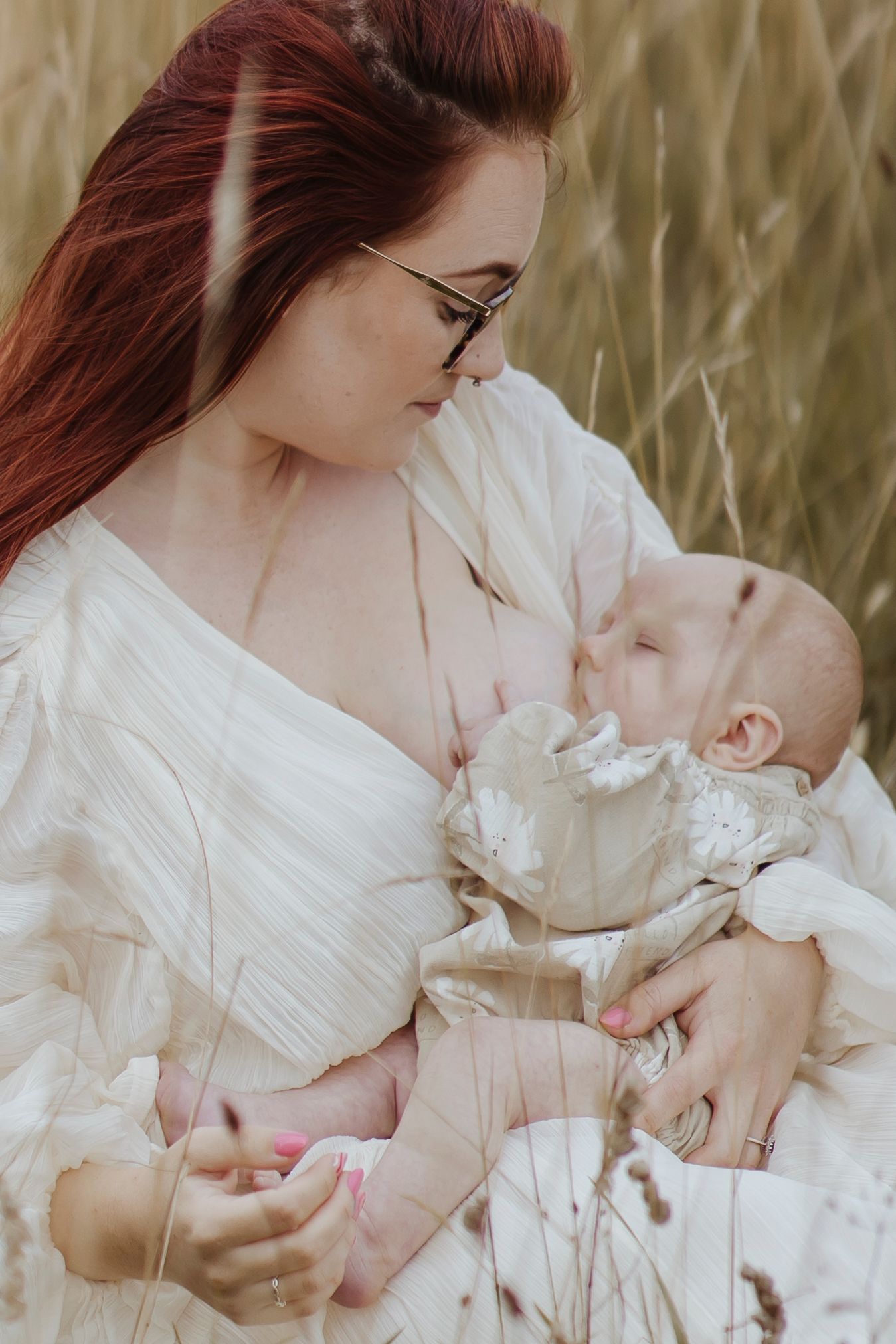 A woman with long red hair breastfeeds a baby while sitting in tall grass, looking down at the child with a calm, tender expression.