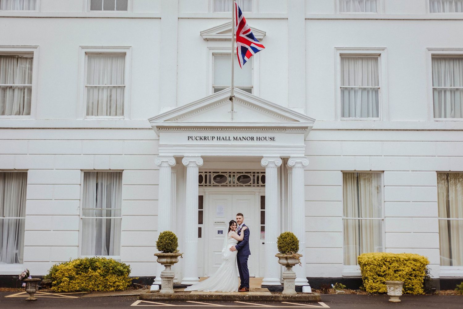 A newlywed couple stands in front of the white, columned Pickering Manor House beneath a flying Union Jack.