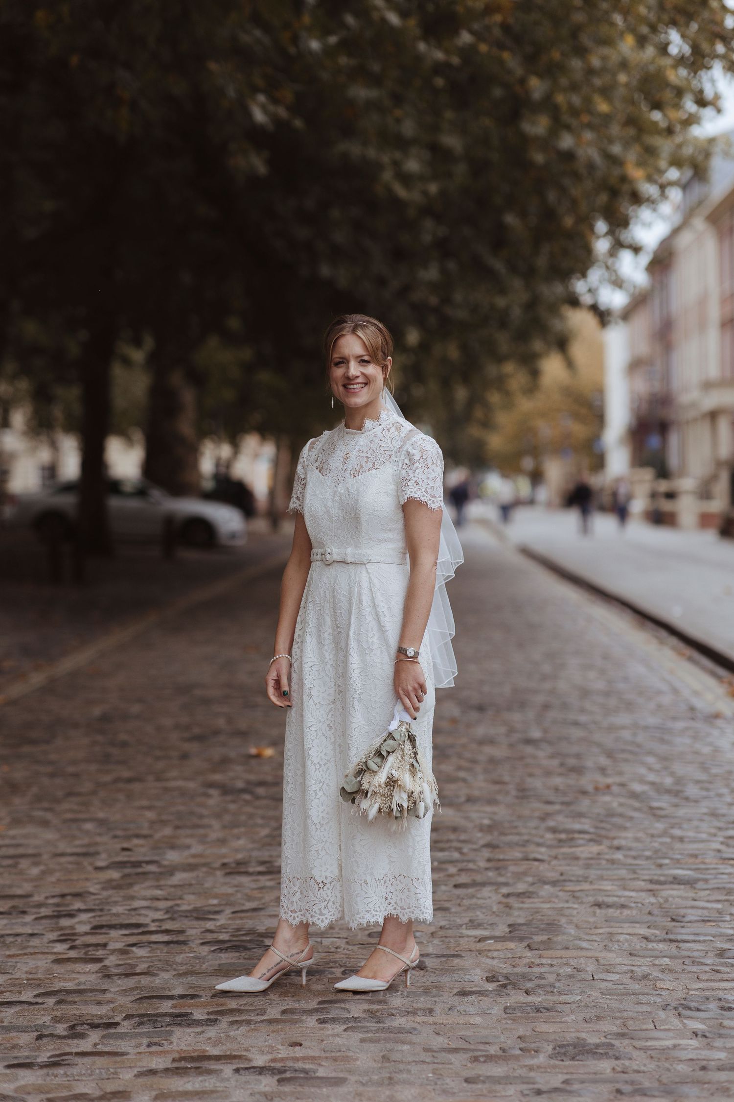 A woman in a white lace dress stands on a cobblestone street holding a small bouquet.