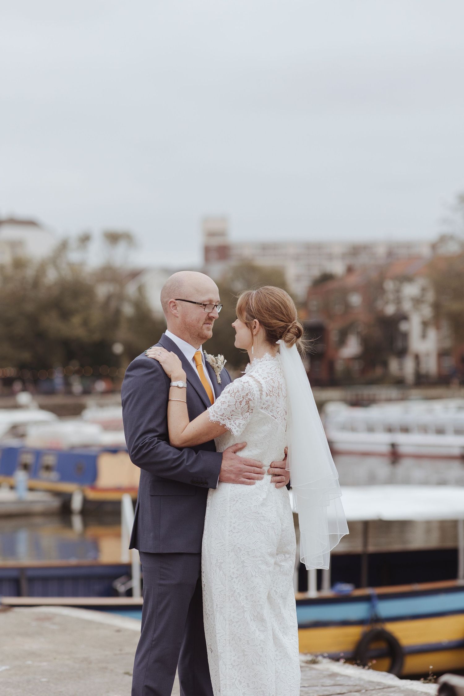 A bride and groom embrace by the waterfront, sharing a quiet, loving moment against a backdrop of boats and soft, overcast sky.