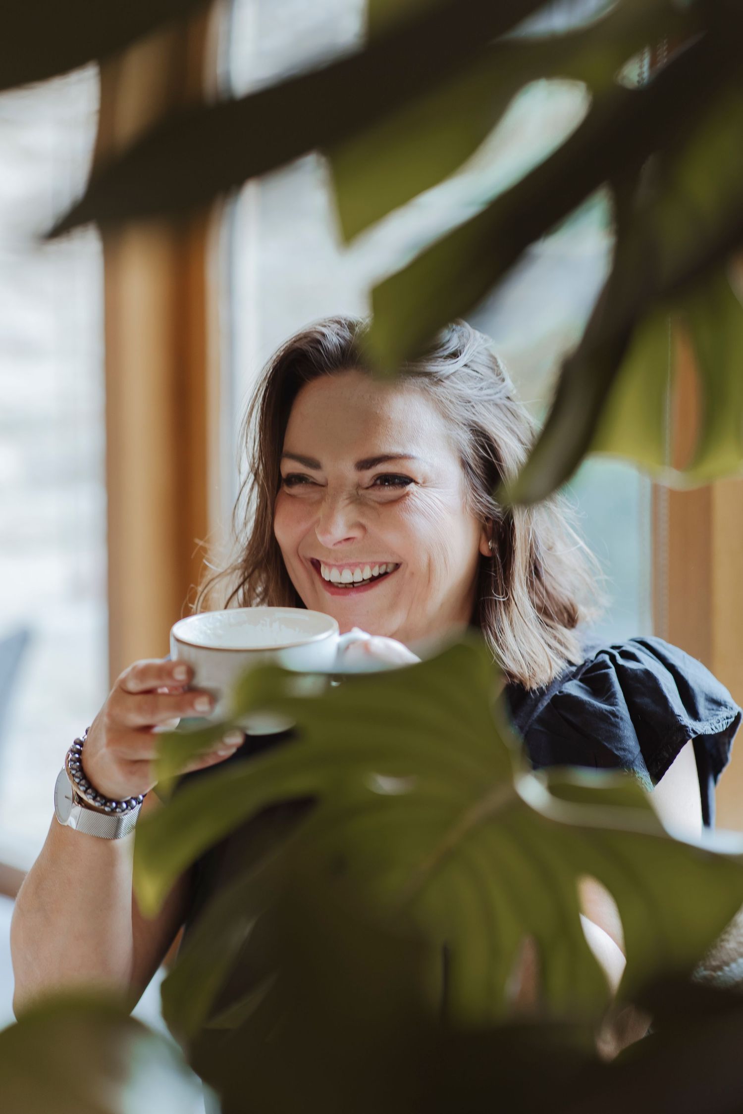 A woman smiles warmly while holding a cup, framed by large green leaves, enjoying a calm moment by a window.