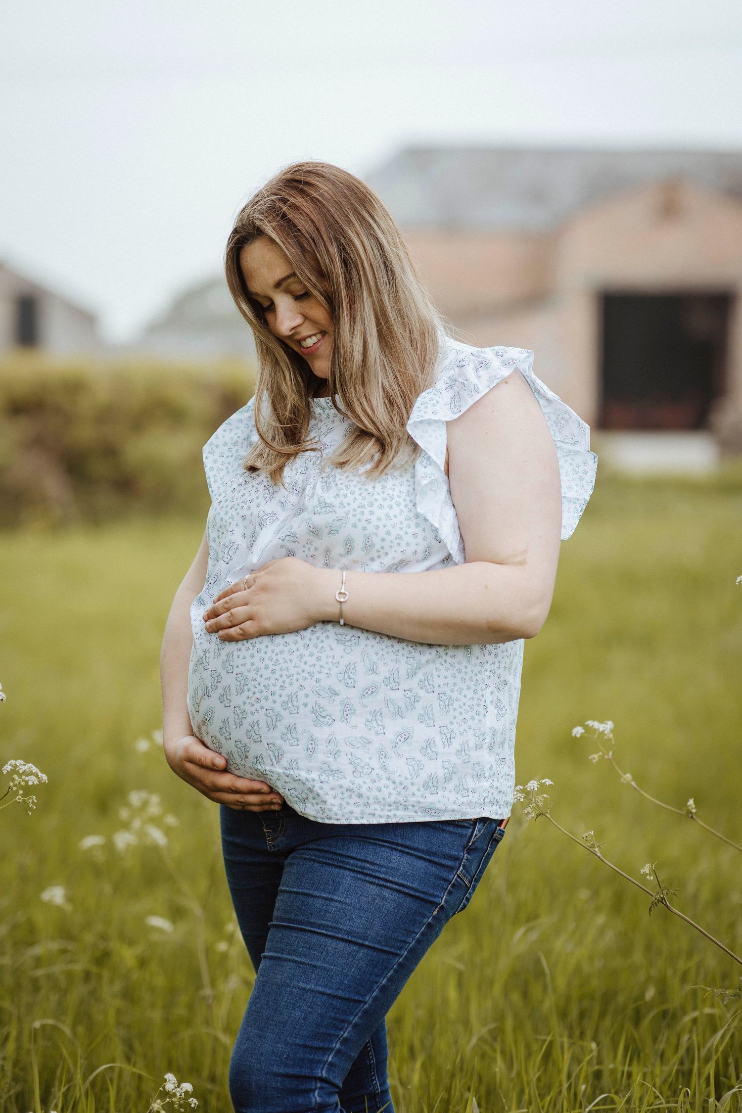 A woman stands in a grassy field, gently holding her pregnant belly with a warm, peaceful smile.