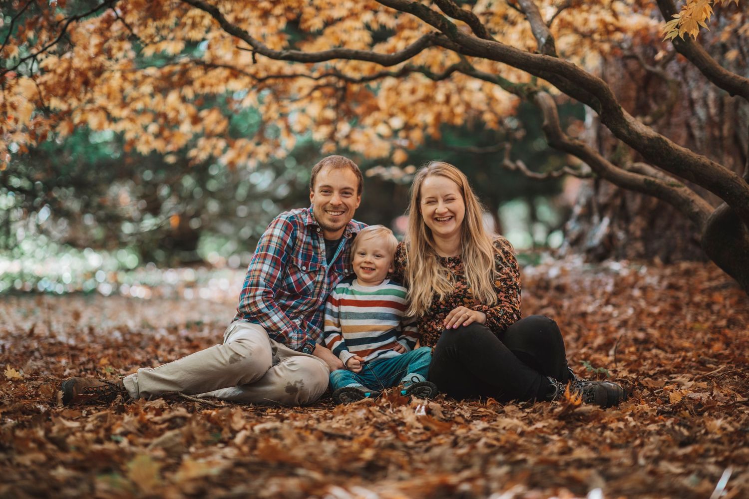 Family sitting together on fallen leaves under autumn trees, smiling at the camera in a warm outdoor setting.