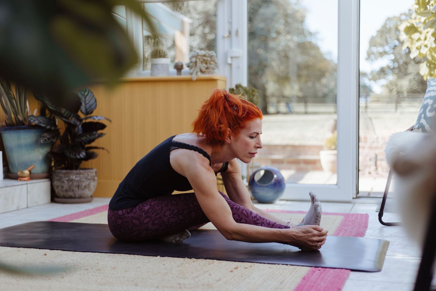 A person practices a deep stretch on a yoga mat in a bright, plant-filled room with sunlight streaming in.