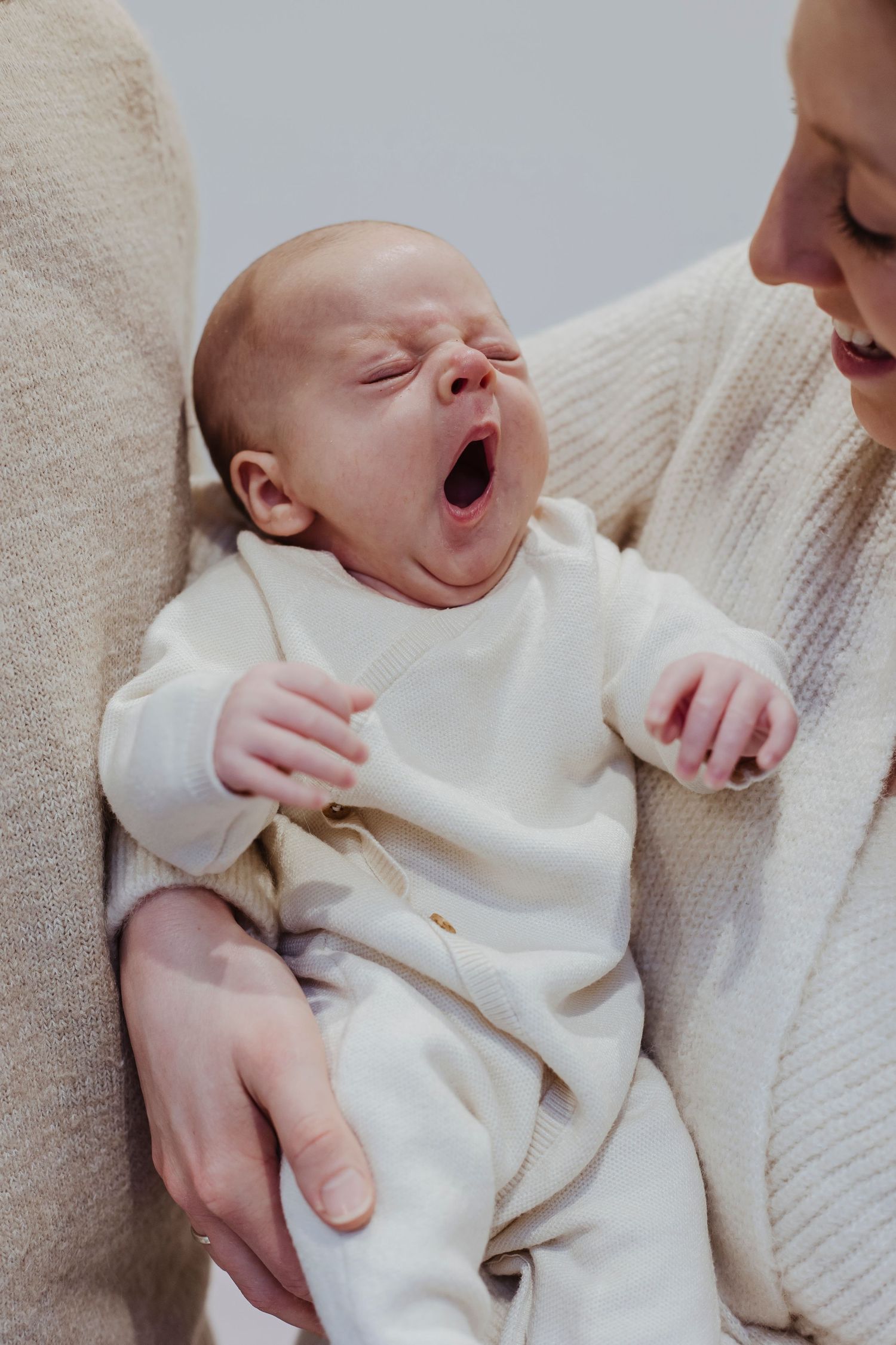A baby yawns wide in a cozy embrace, held close and comforted in soft, warm tones.