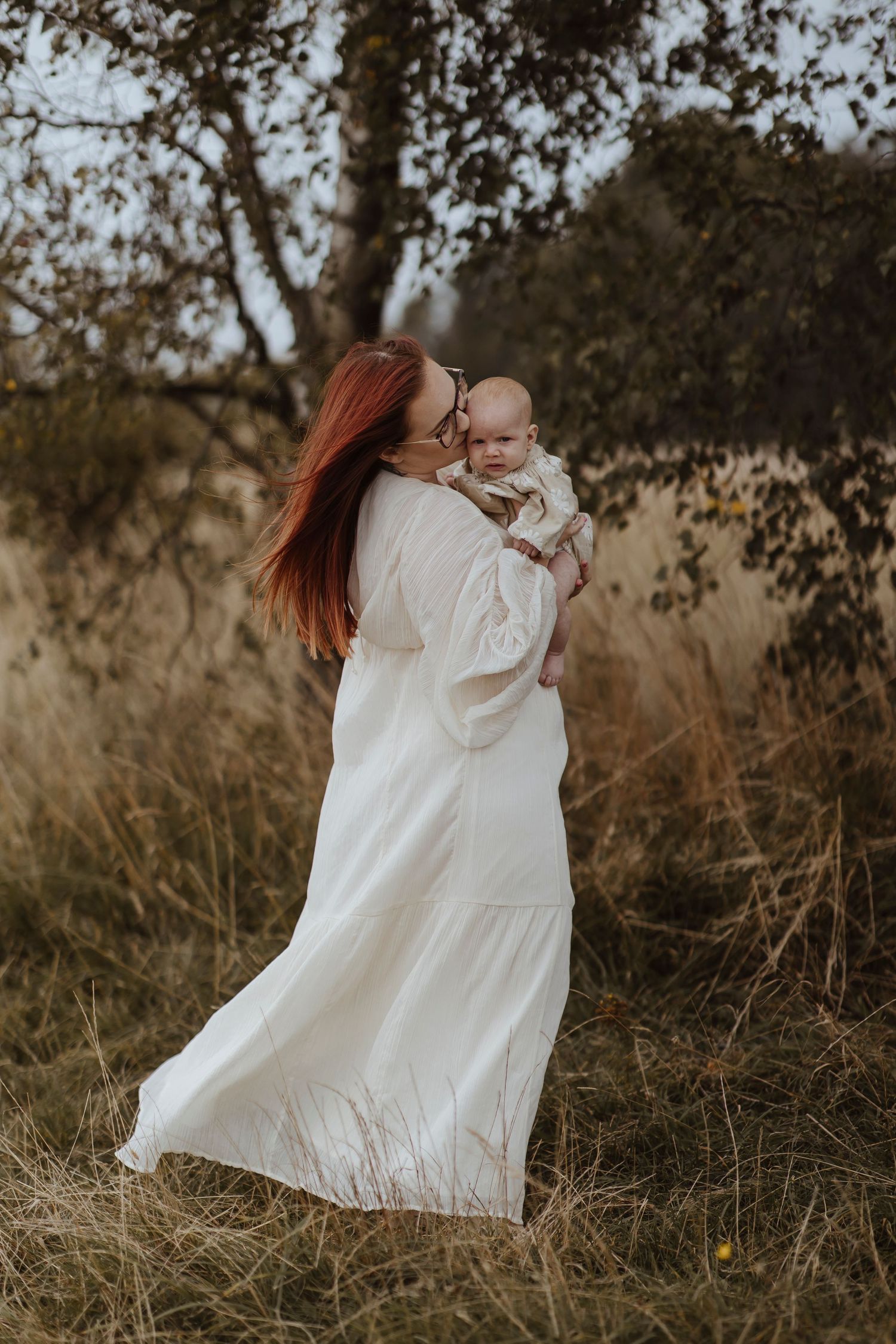 A woman in a flowing white dress walks through tall grass while holding a baby, both looking relaxed in the natural, breezy se