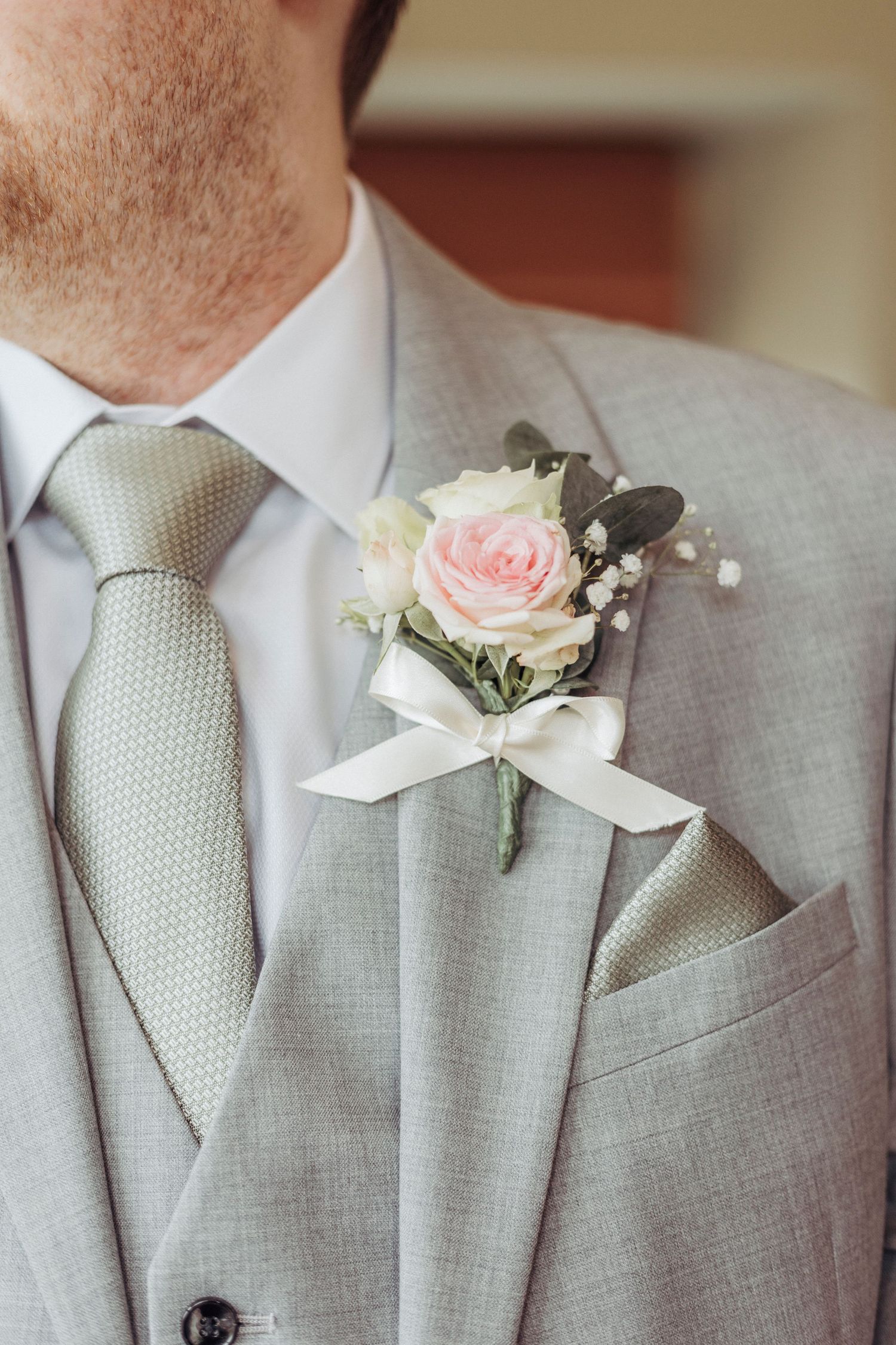 A close-up of a groom’s elegant boutonnière—soft pink rose, greenery, and a white ribbon—pinned to a light grey suit.