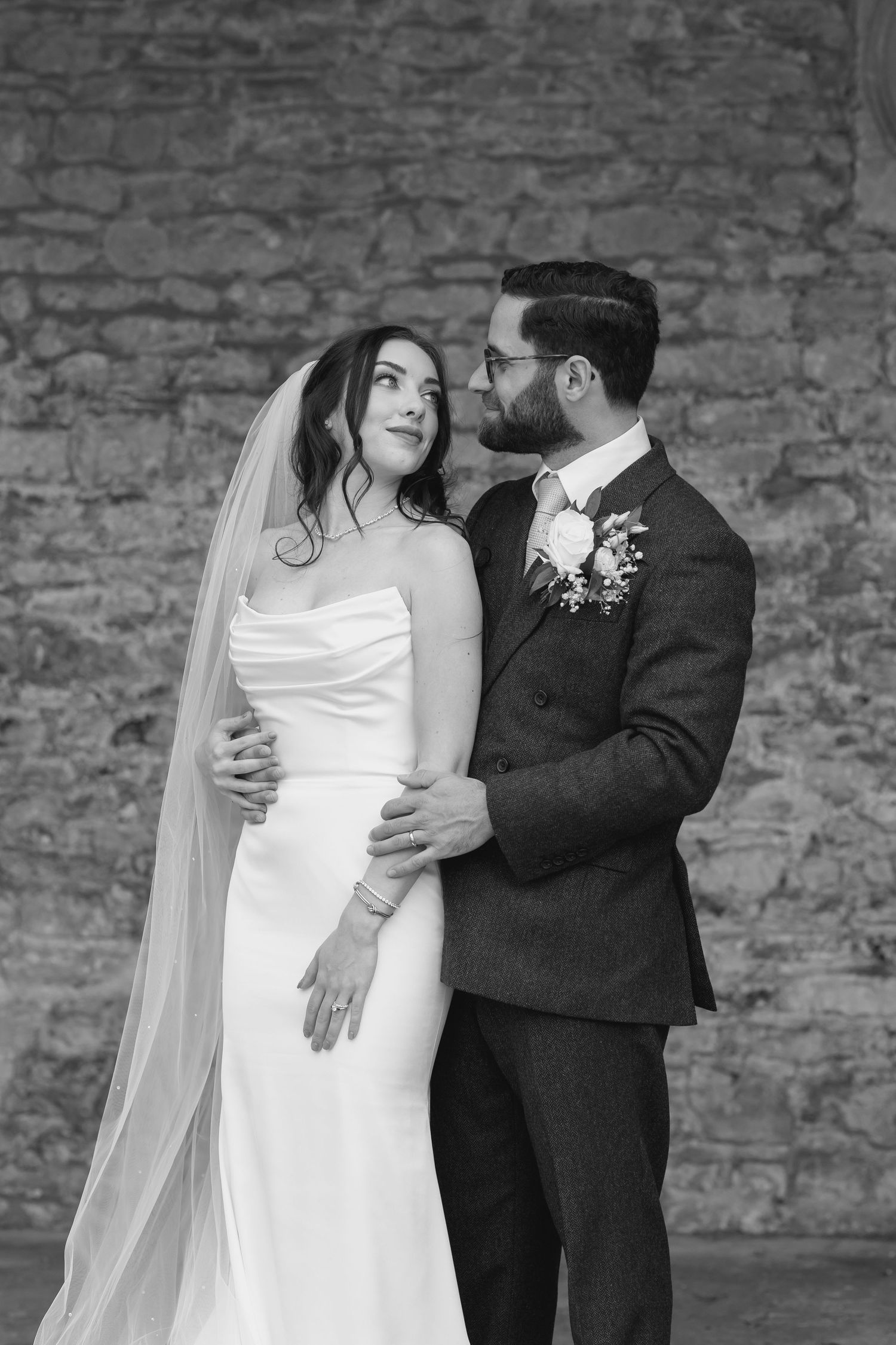 bride and groom standing in front of a stone wall glancing at each other