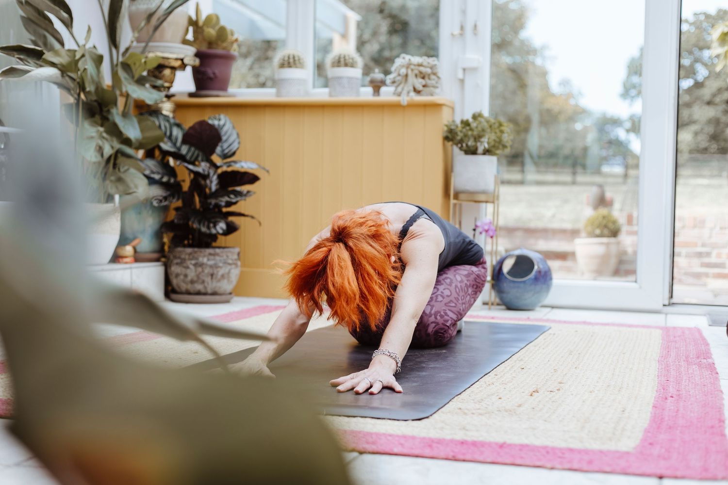 A person with bright hair stretches forward on a yoga mat in a cozy, plant-filled room, reaching out with both hands in a relaxed pose.