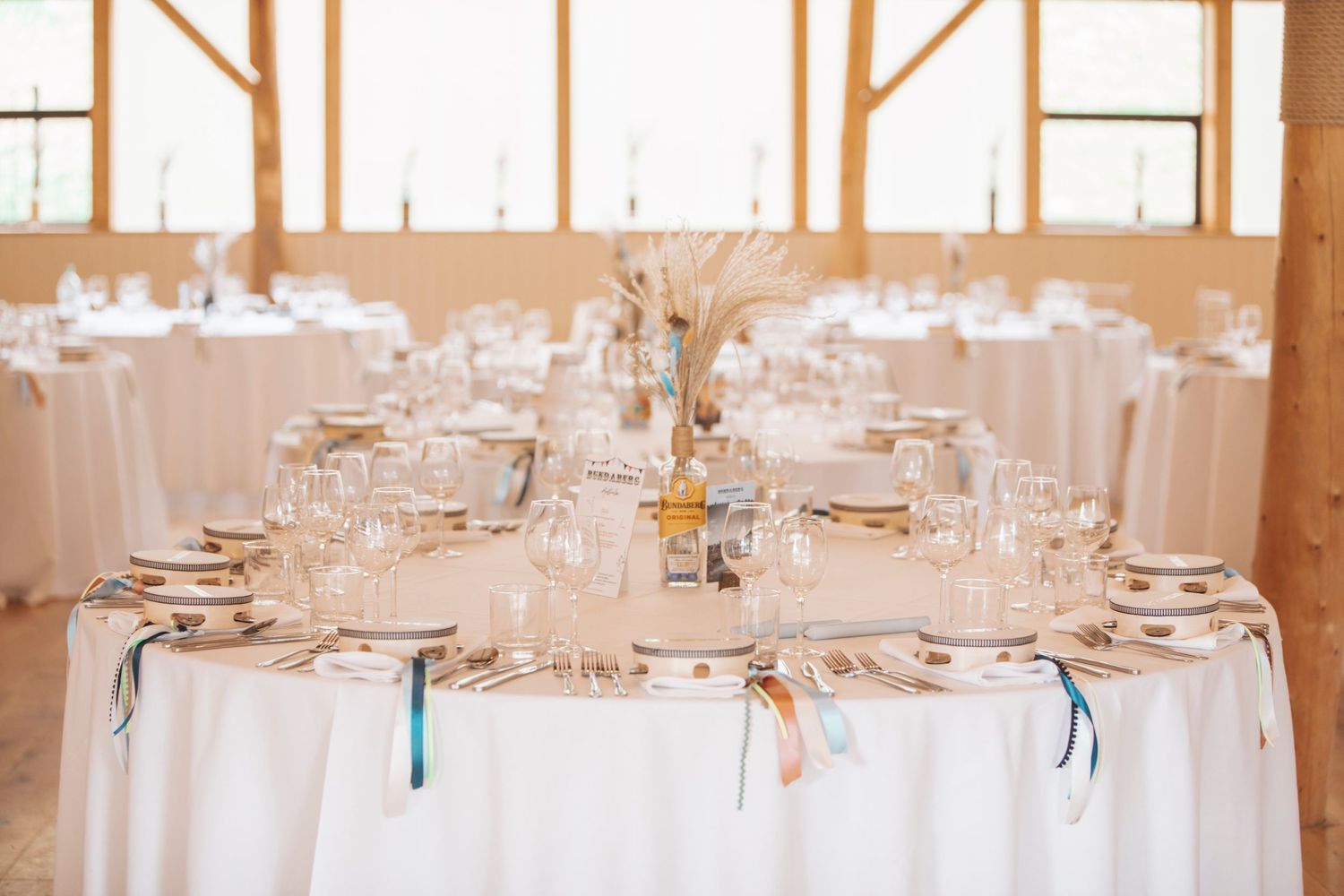 Elegant wedding reception table set with white linens, glassware, plates, and a simple centerpiece inside a bright rustic venue.