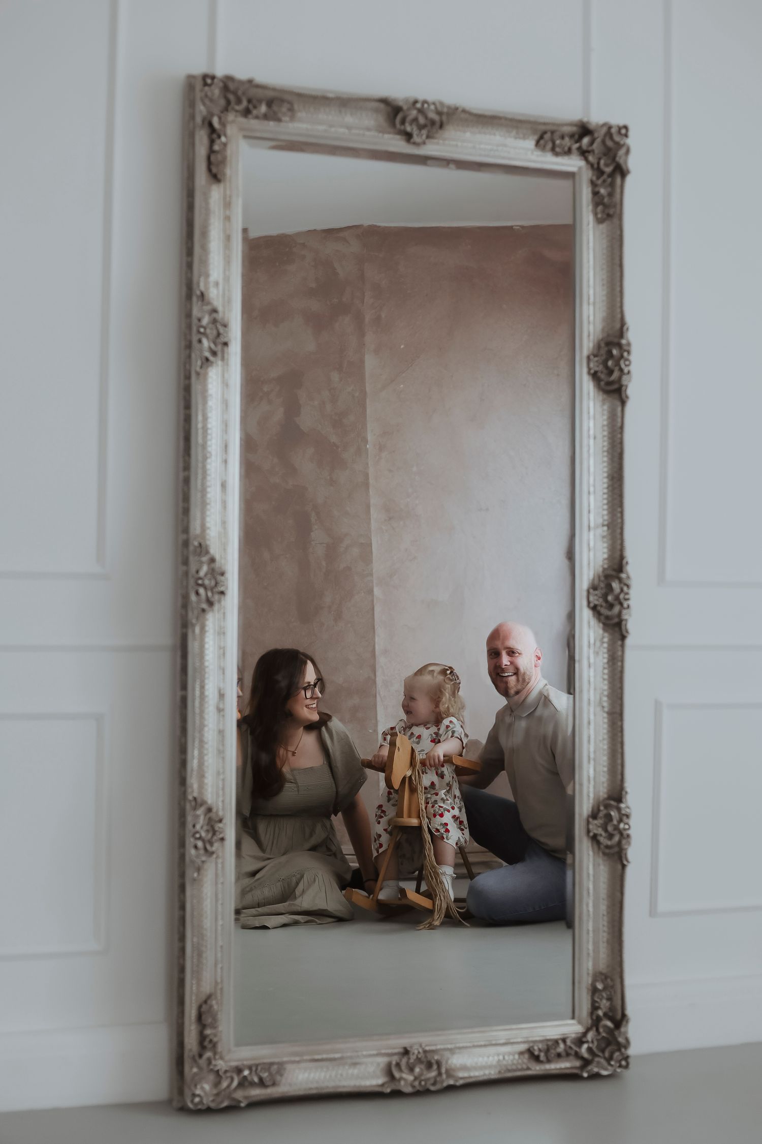 Family sitting together in a studio reflected through an ornate vintage mirror, with parents smiling at their toddler holding a wooden toy.
