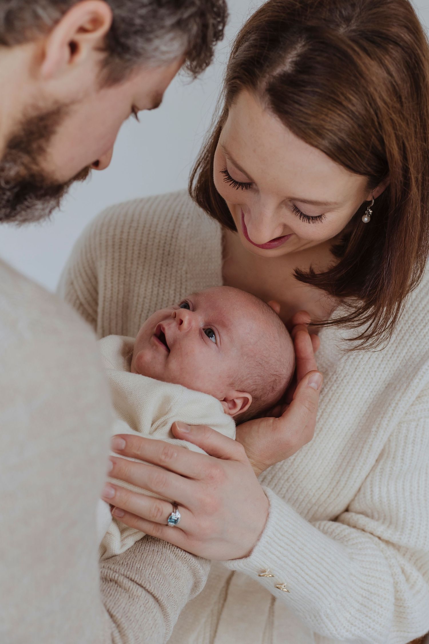 A newborn gazes up as two adults lean close, sharing a quiet, tender moment together.