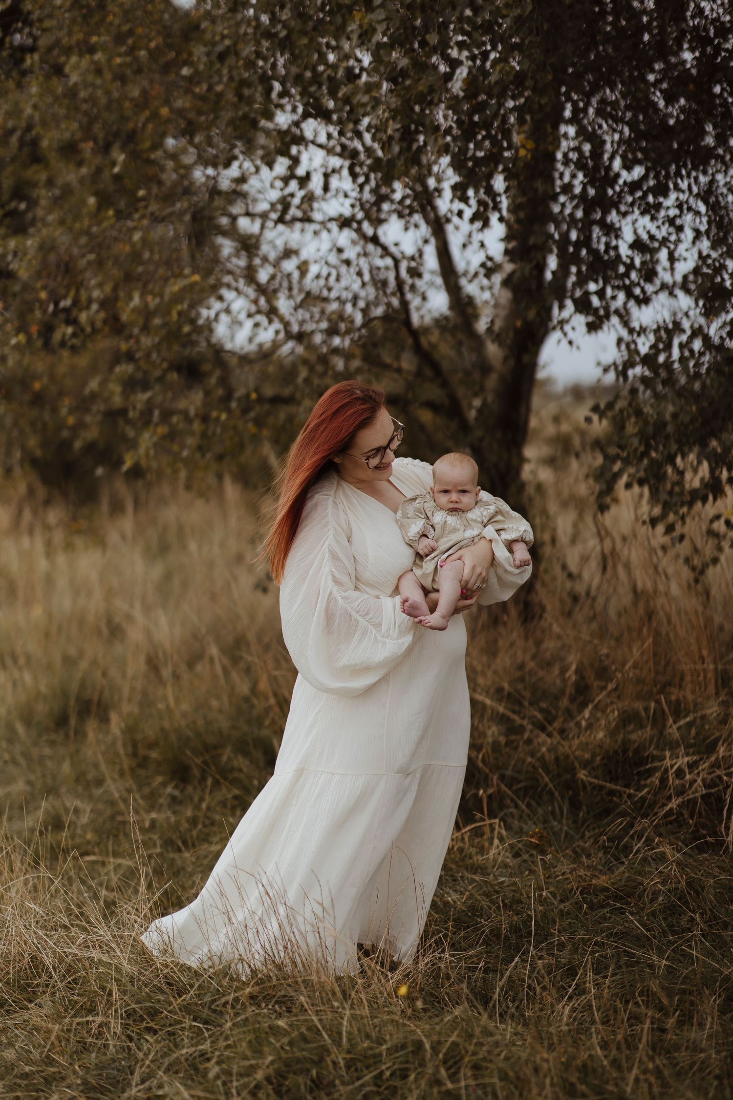 A woman in a long white dress holds a baby while standing in a grassy field, surrounded by trees and soft, natural light.