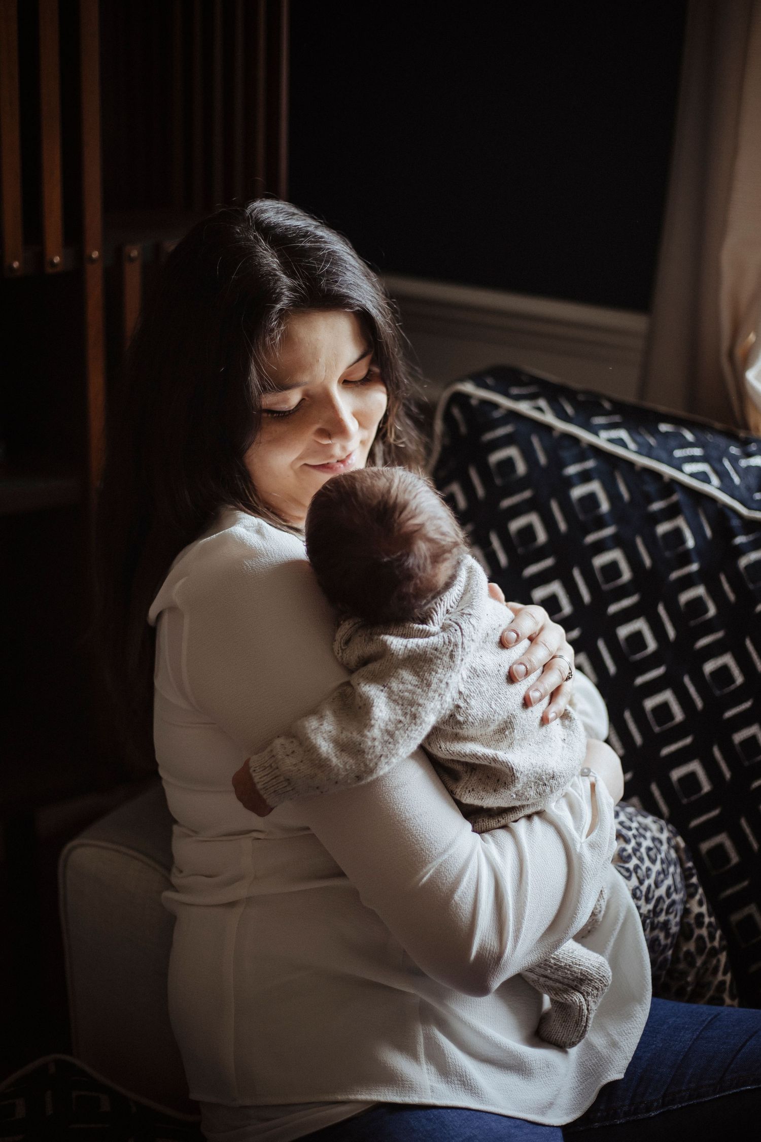 A woman sits by a window, gently holding a newborn against her shoulder, both resting peacefully.