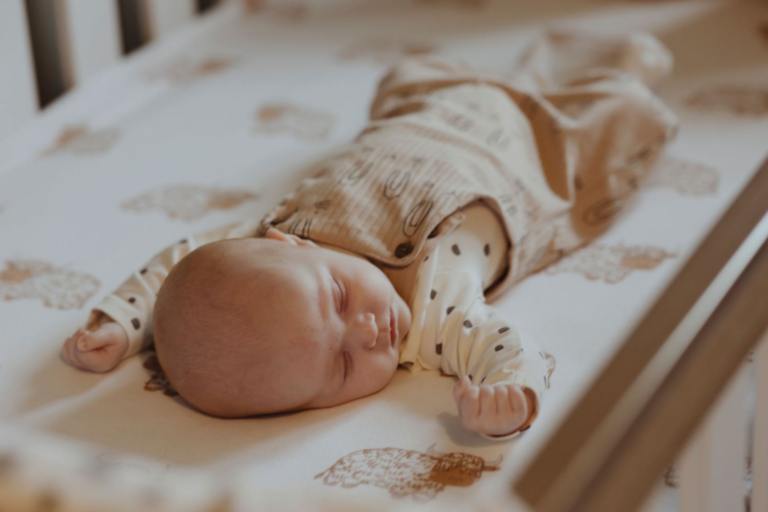 A baby is lying on their back in a crib, arms stretched outward, appearing peacefully asleep on a patterned sheet.
