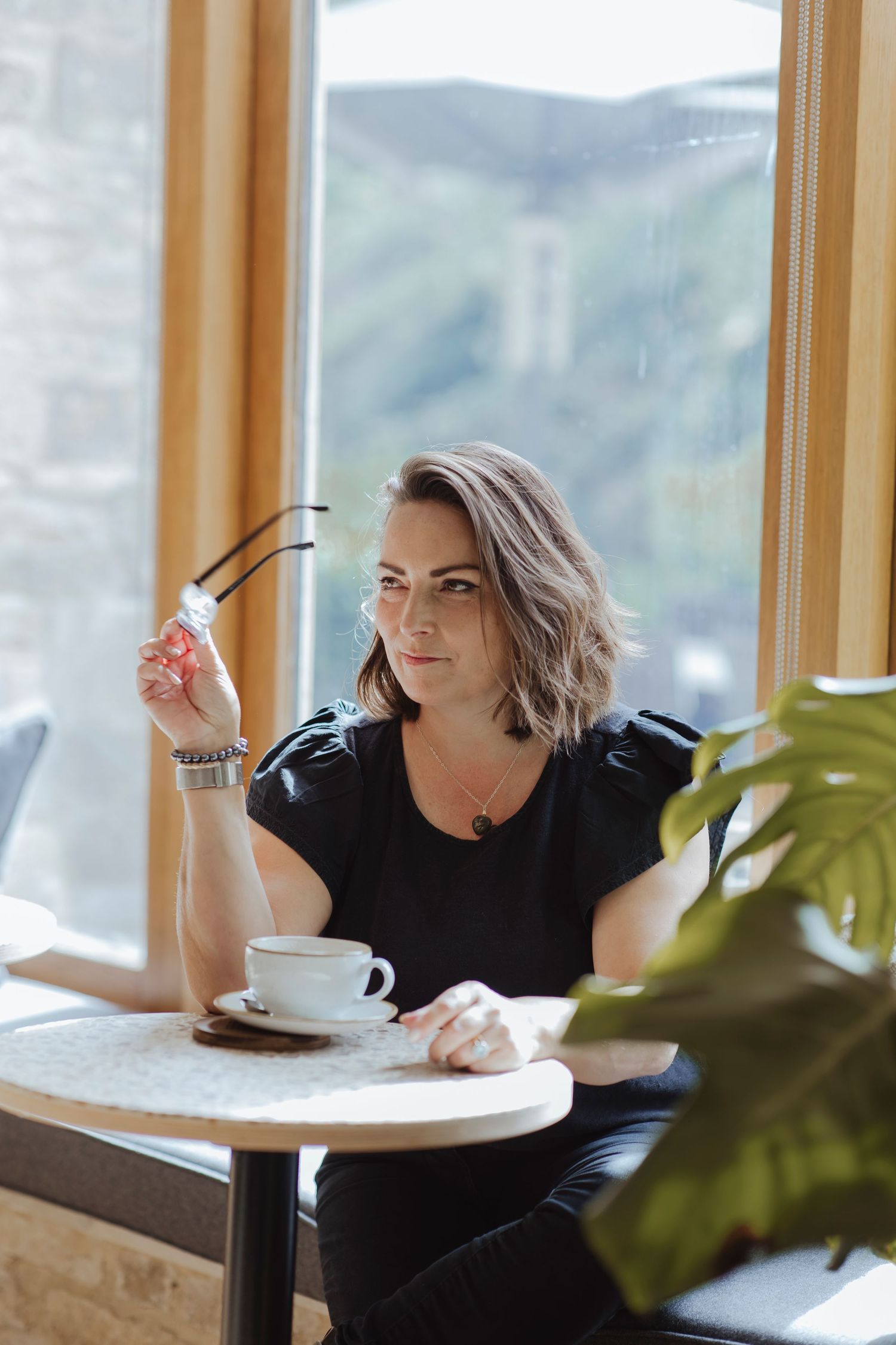 A woman sits by a bright window with a cup of coffee, holding her glasses thoughtfully as she gazes outside.