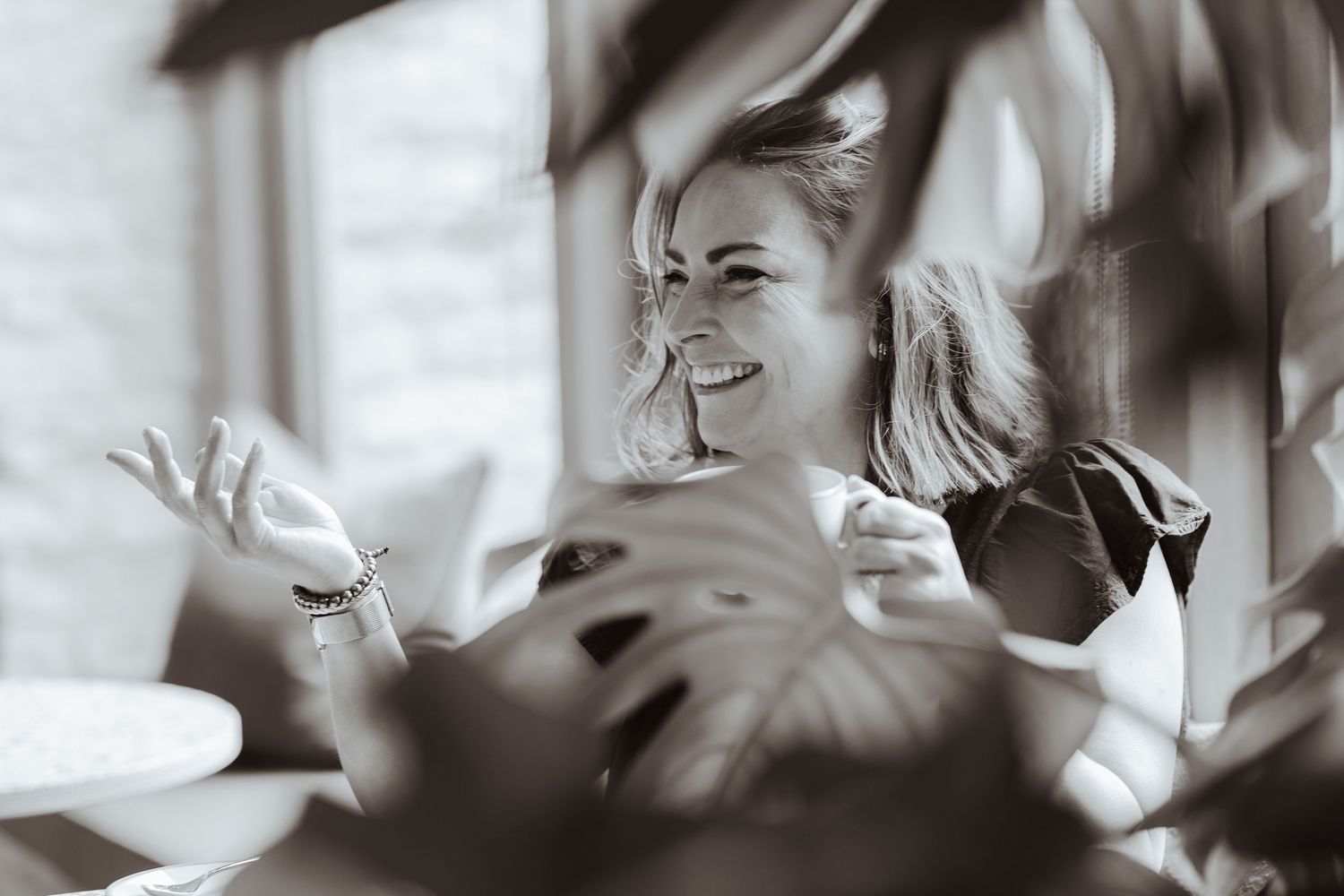 A person smiles and gestures mid-conversation, partially framed by large leaves, creating a warm and candid moment.